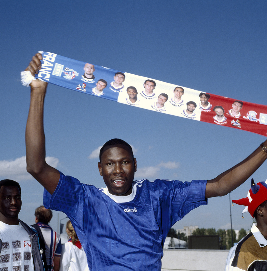 A man in a blue shirt holds up a France soccer scarf featuring players’ photos. He is smiling with arms raised outdoors, with other people and a building in the background.