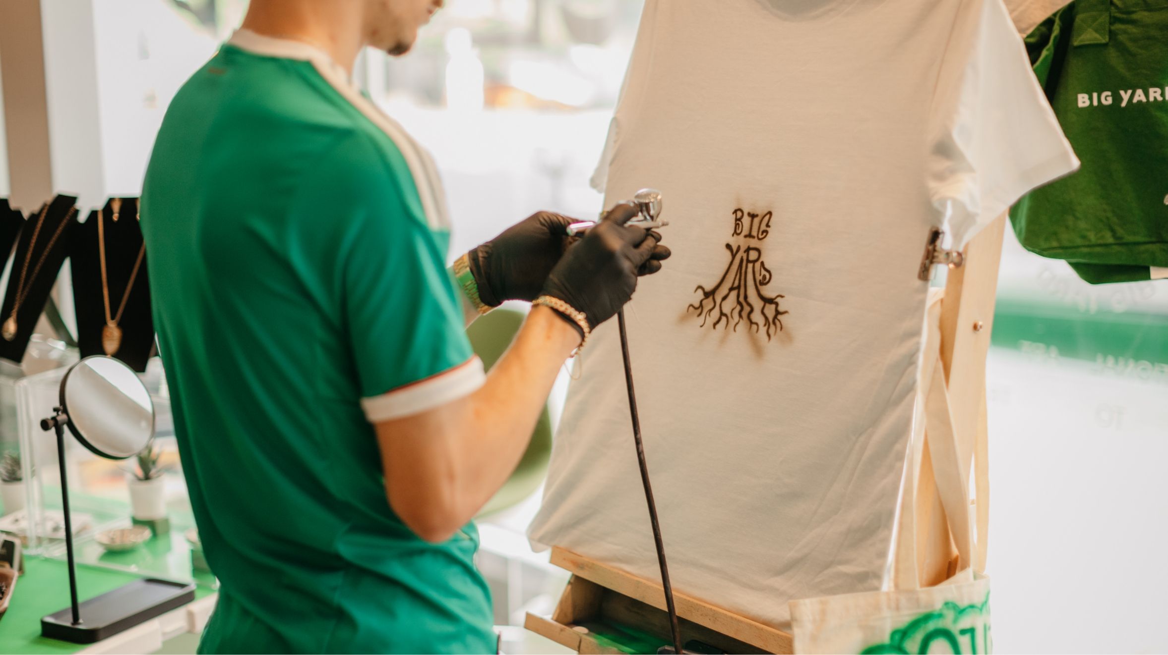 A person wearing a green shirt airbrushes a design onto a white T-shirt. The T-shirt is displayed on a wooden easel.