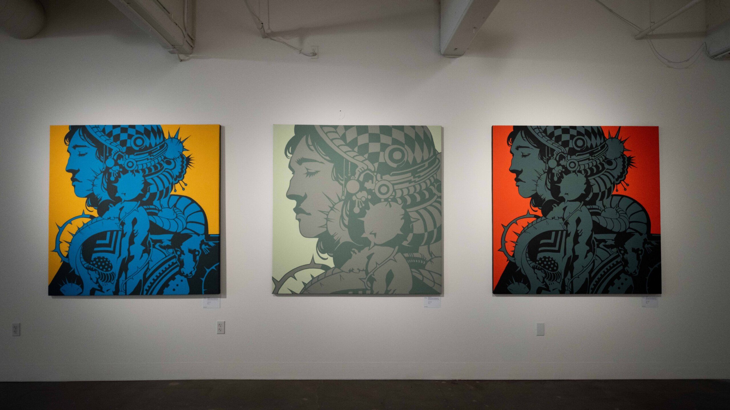 Three colorful canvas artworks depicting stylized profiles of a person with intricate patterns are displayed on a white gallery wall. The backgrounds are blue, green, and orange.