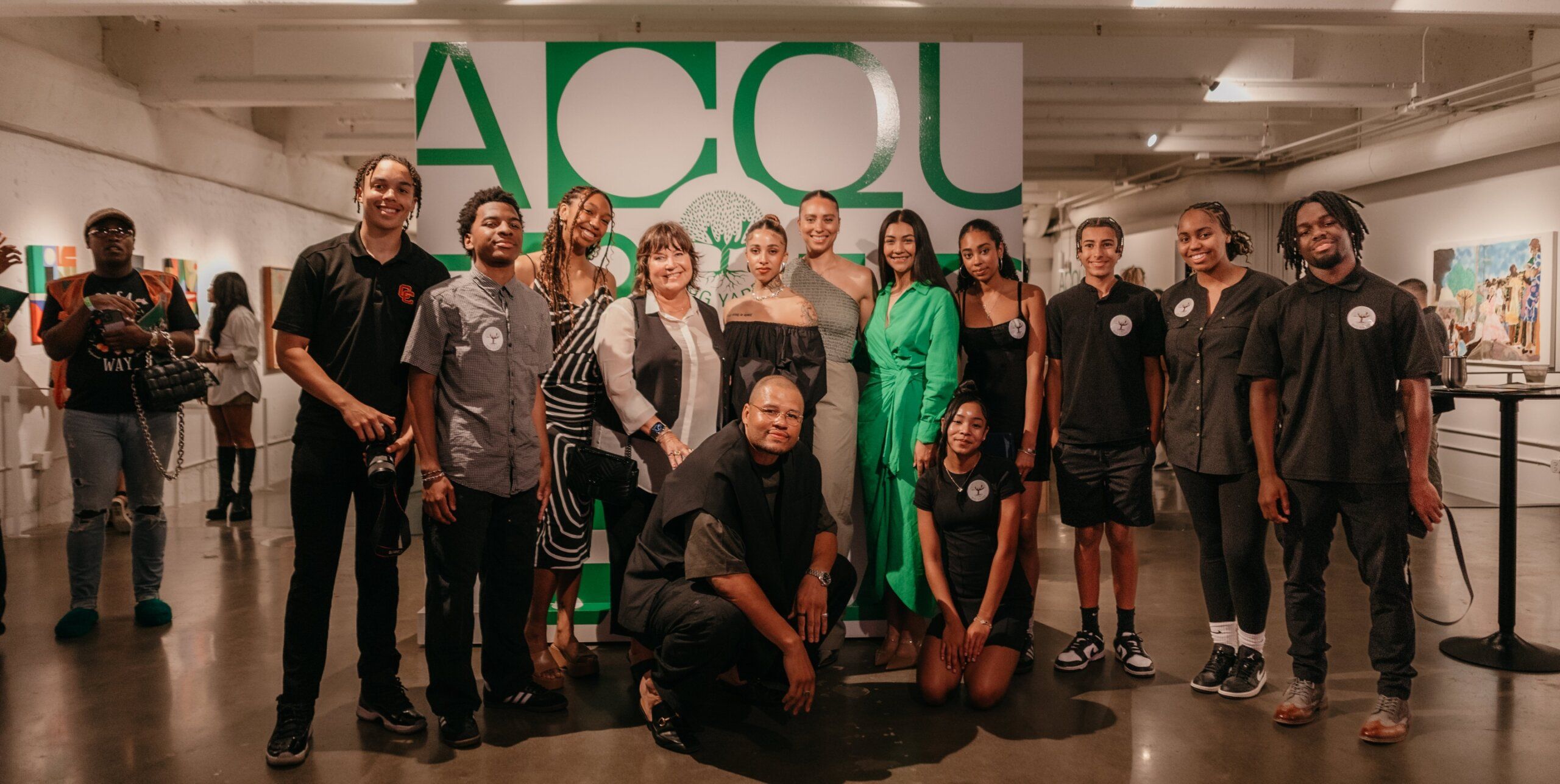 A group of people pose together in front of a display with large green letters "ACQUA" at an indoor event. Some are dressed in casual attire, while others wear more formal clothing.