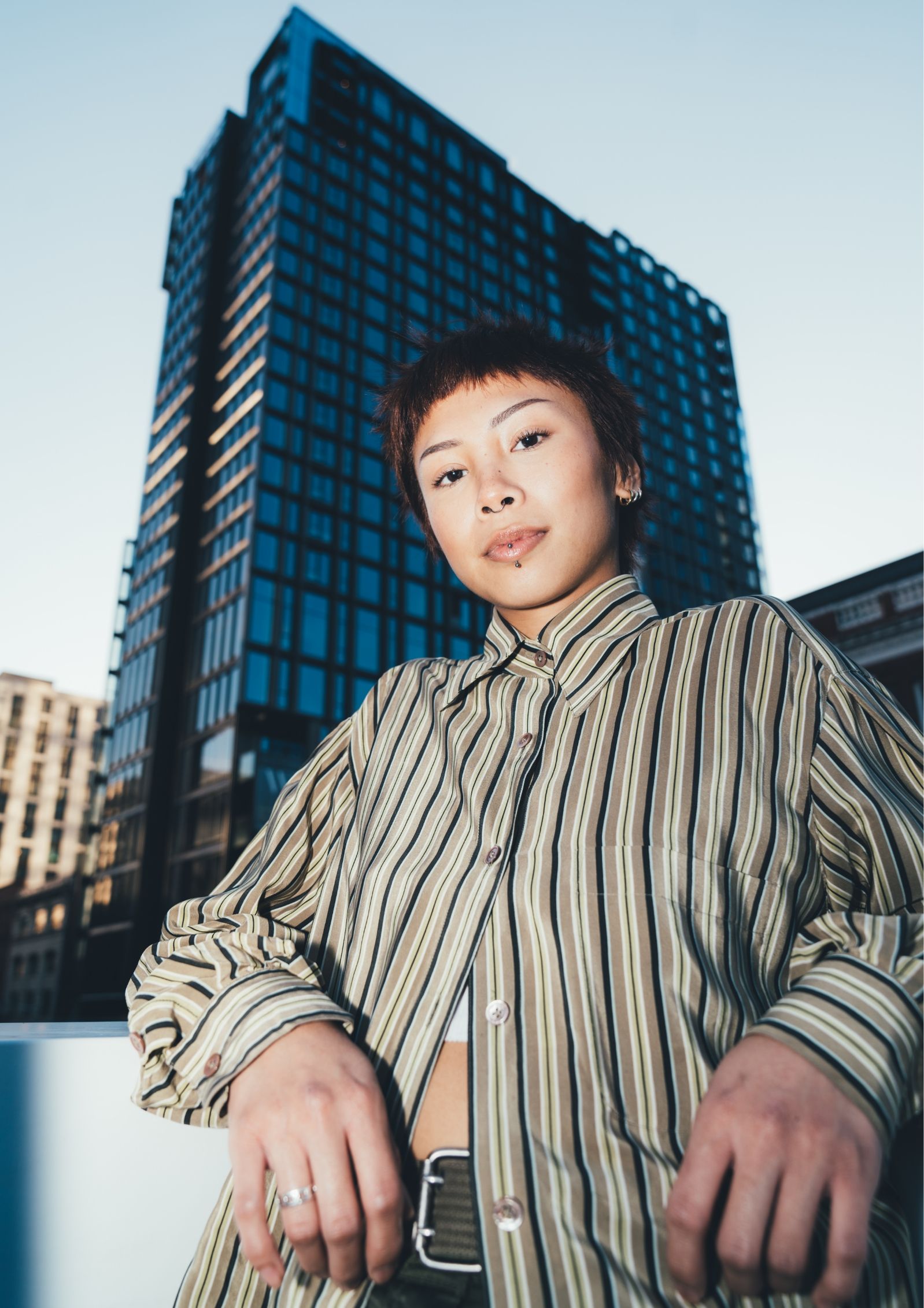 A woman in a striped shirt stands confidently with a high-rise building in the background under a clear sky.