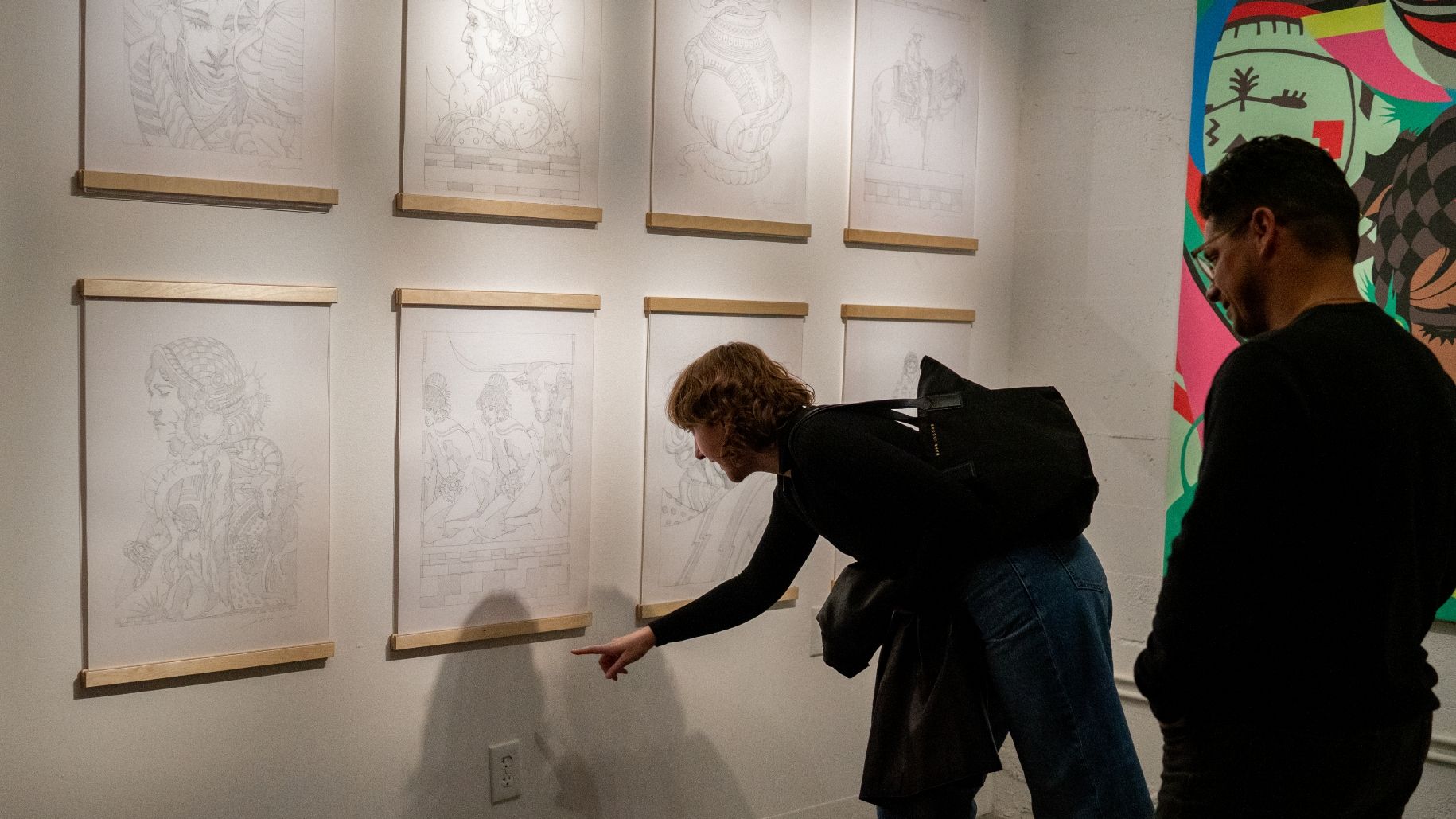 Two people observing and discussing framed pencil sketches displayed on a gallery wall, with one person pointing at a specific sketch.