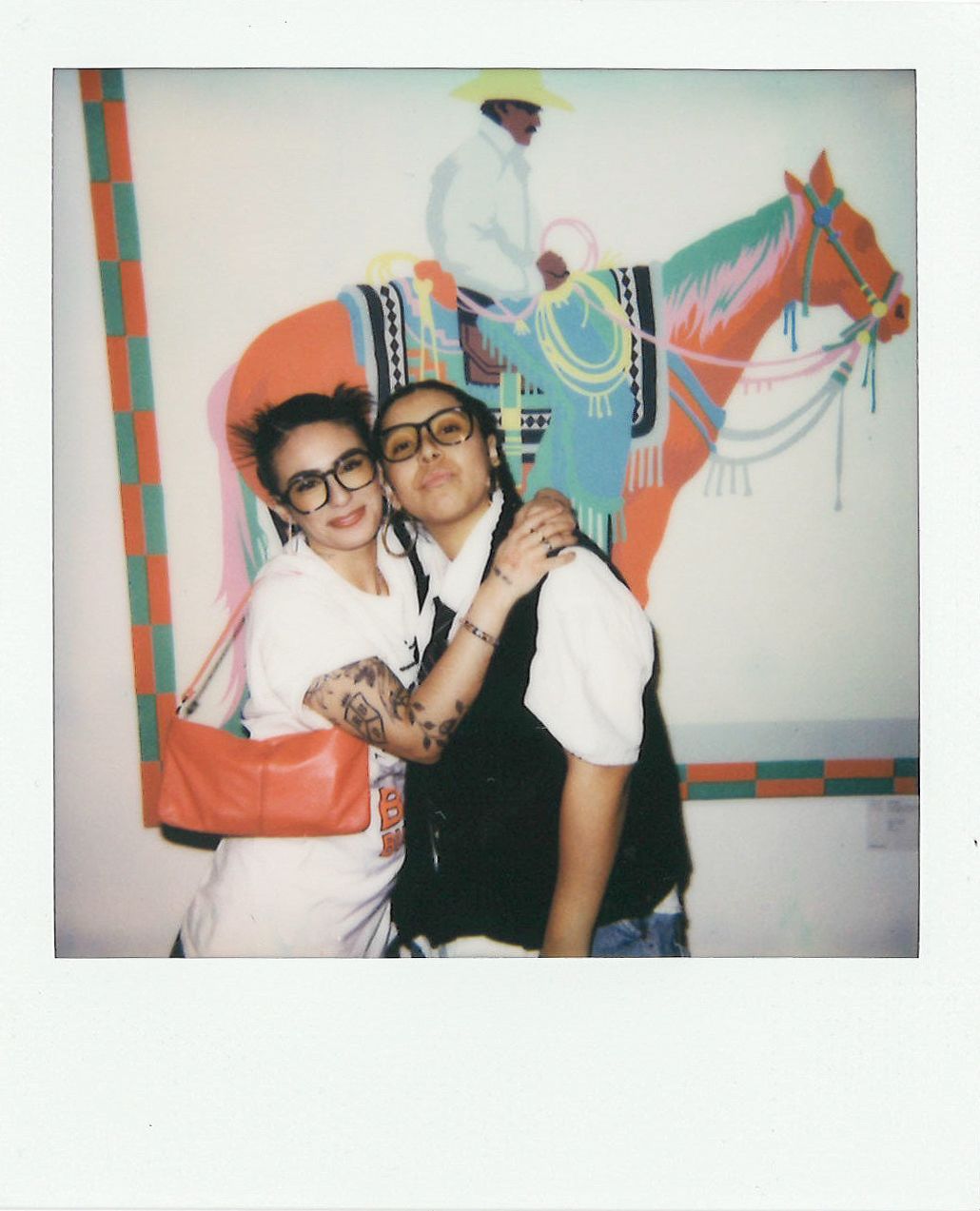 Two individuals pose closely together in front of a vibrant painting of a person riding a decorated horse. One person wears glasses and holds a red purse, while the other dons a vest and braided hair.