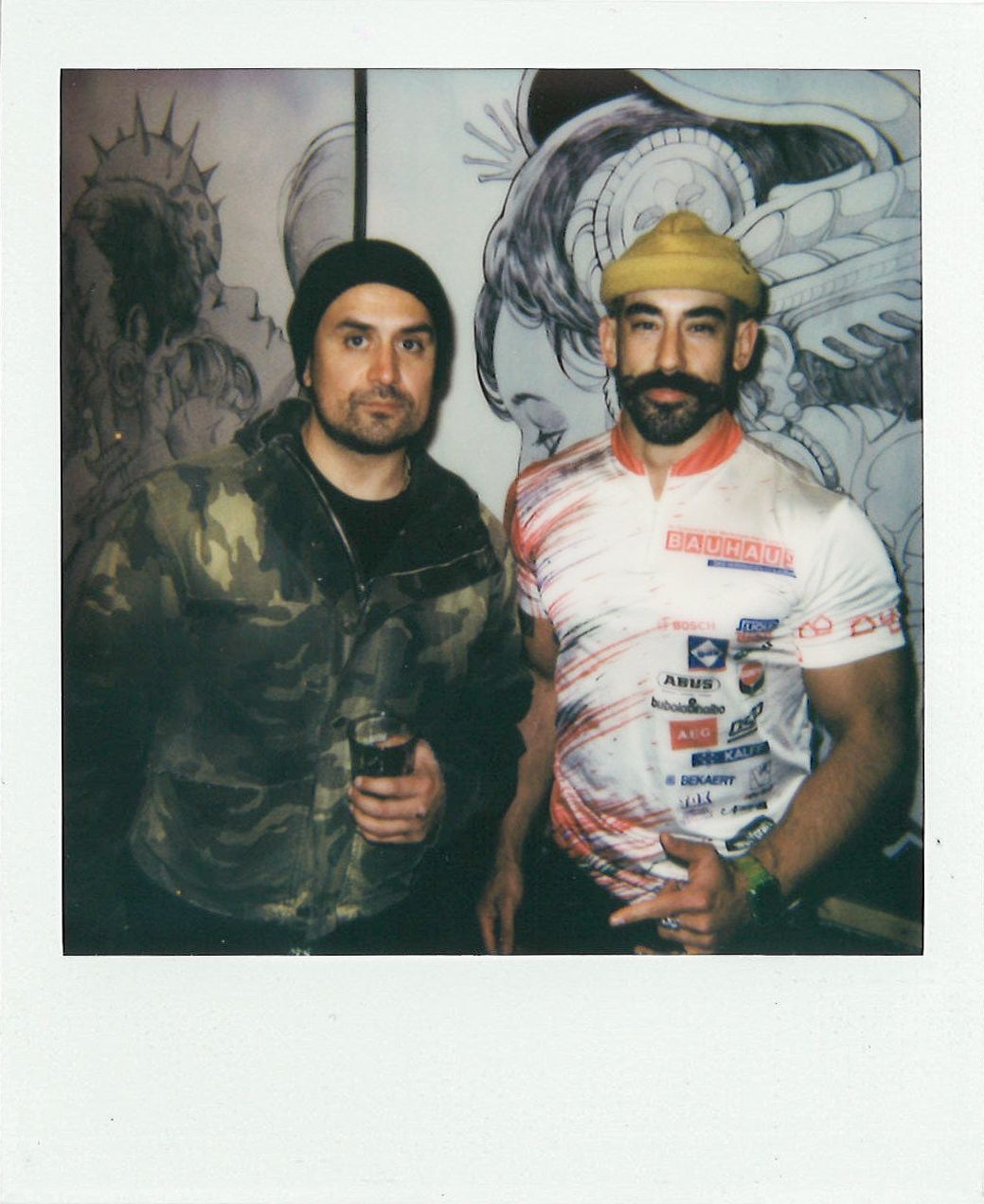 Two men pose for a photo in front of art. One wears a camouflage jacket and a black beanie, and the other wears a yellow cap and a white and red printed shirt.