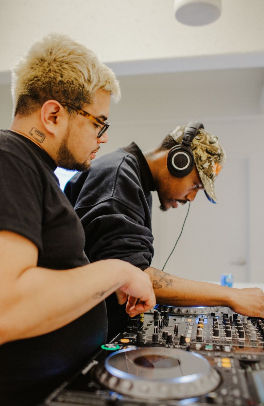 Two individuals are DJing. One is operating the mixer, while the other, wearing headphones, adjusts settings on the equipment.