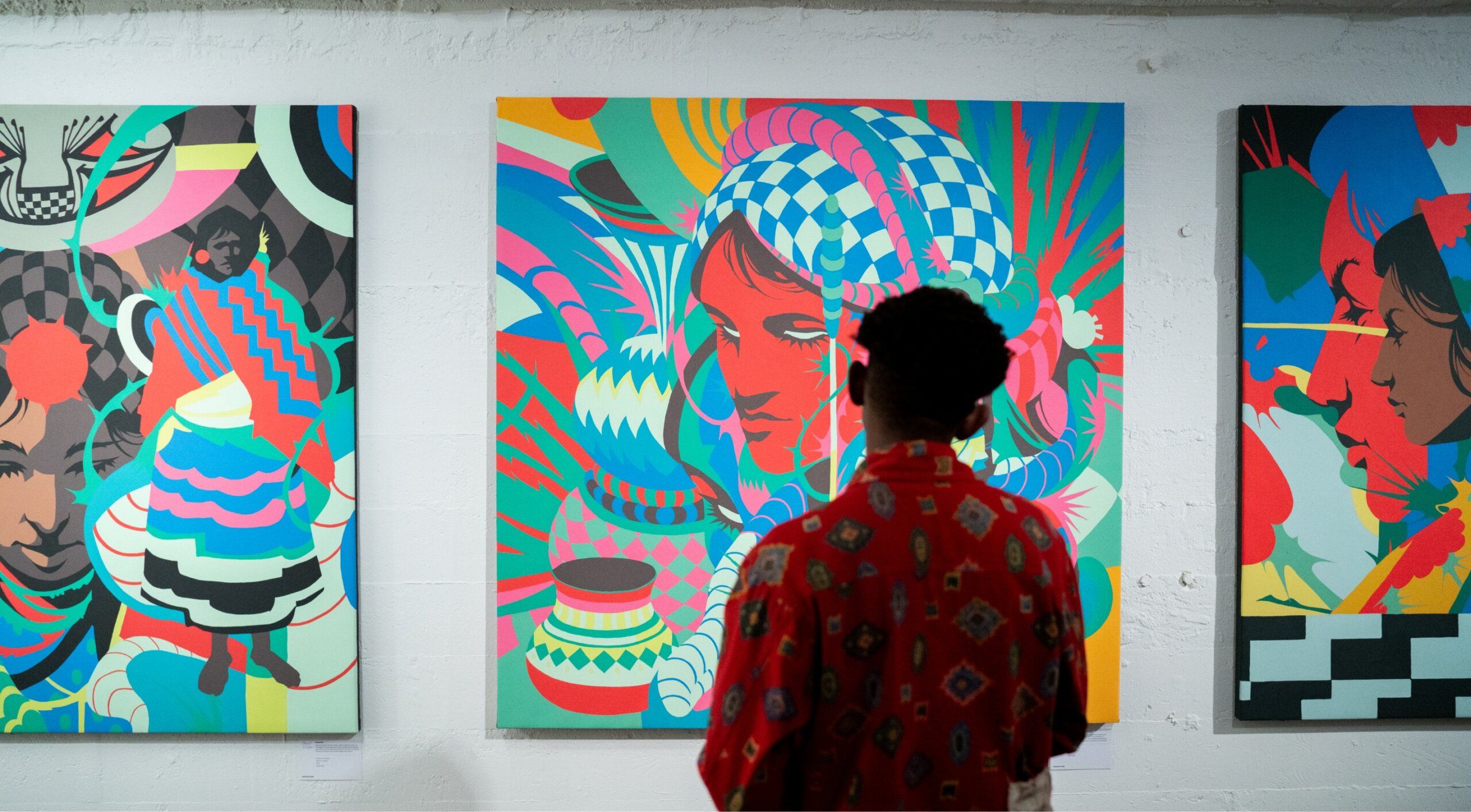 A person in a red patterned shirt views three colorful abstract paintings in an art gallery.