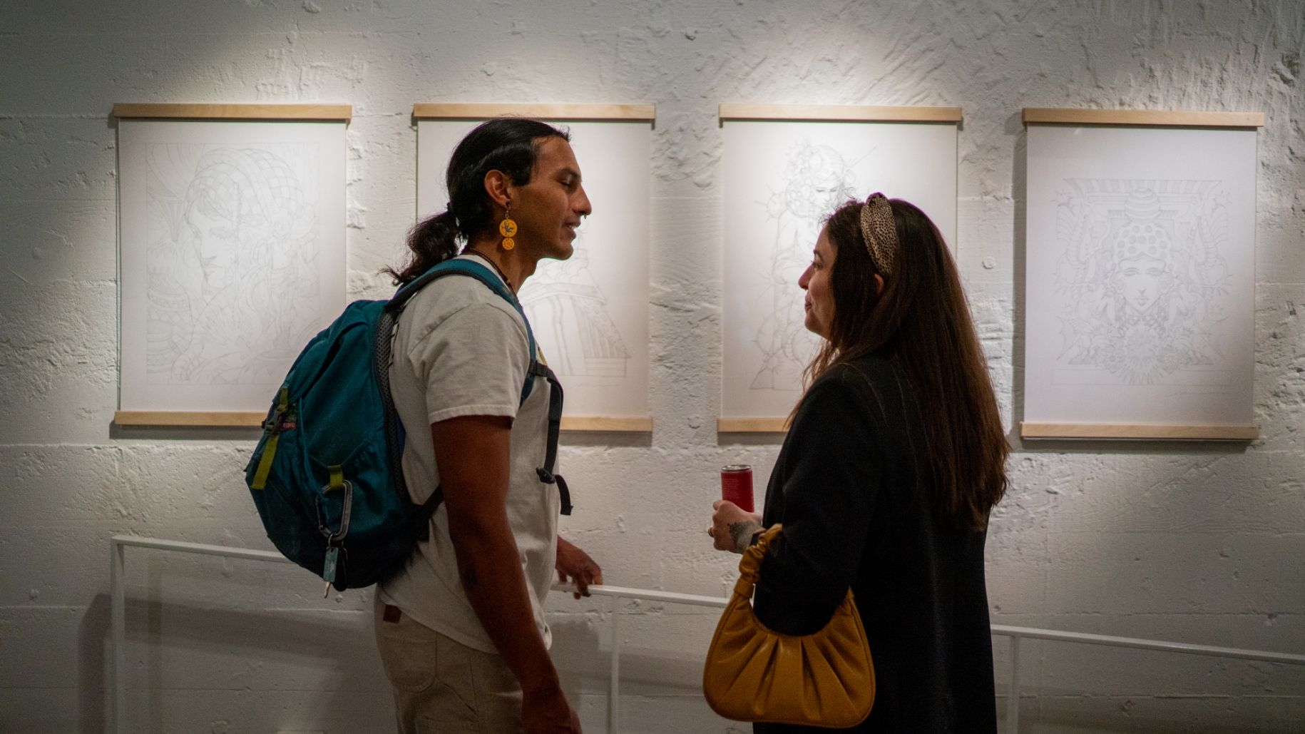 Two people stand in an art gallery discussing framed sketches on a white wall. One holds a drink and the other wears a backpack.