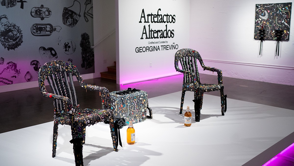 Two chairs and a box covered in various embellishments, with two bottles placed underneath, displayed in an art exhibit titled "Artefactos Alterados" by Georgina Treviño.