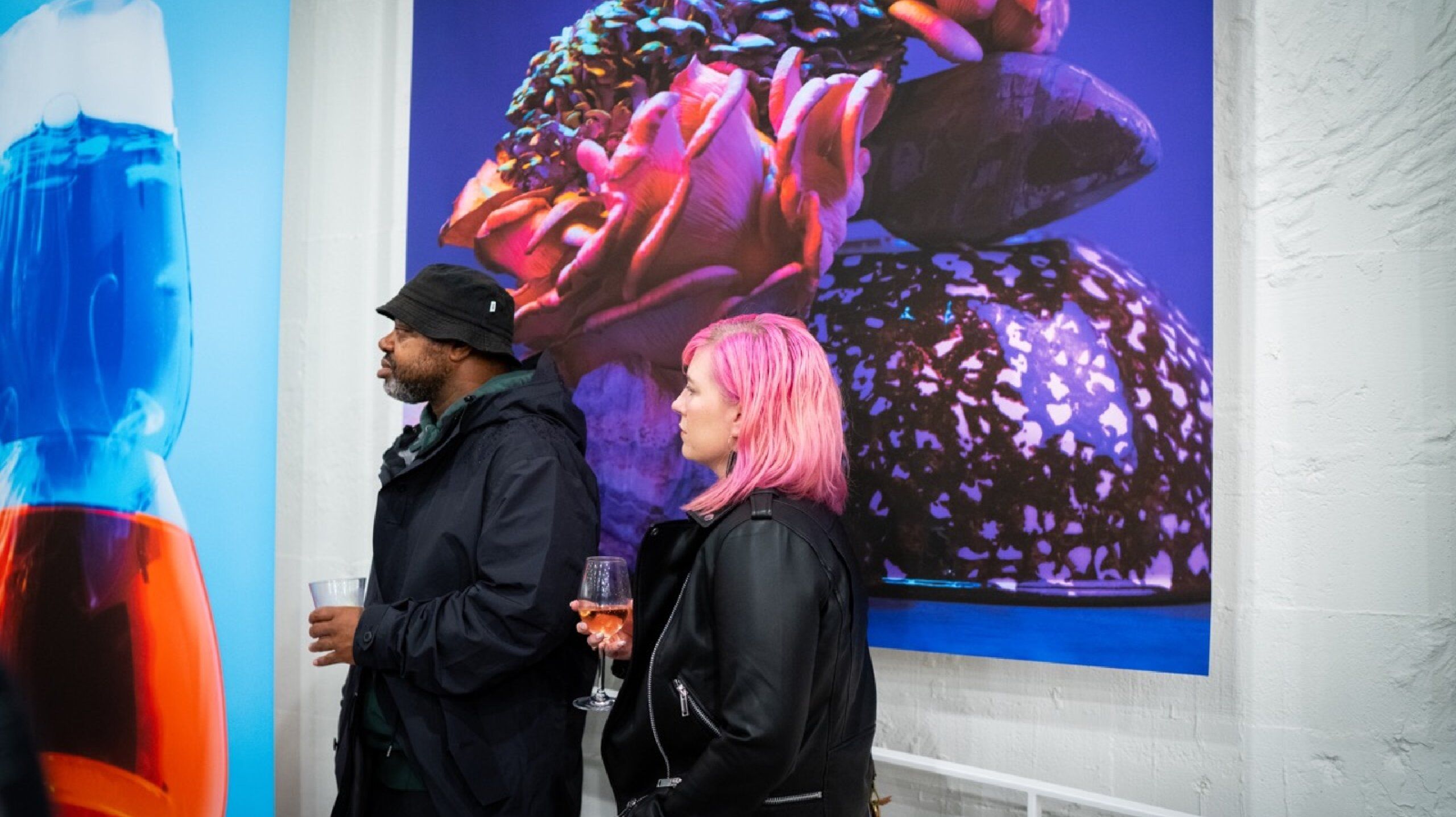 Man and woman with pink hair stand in front of print listening to speakers