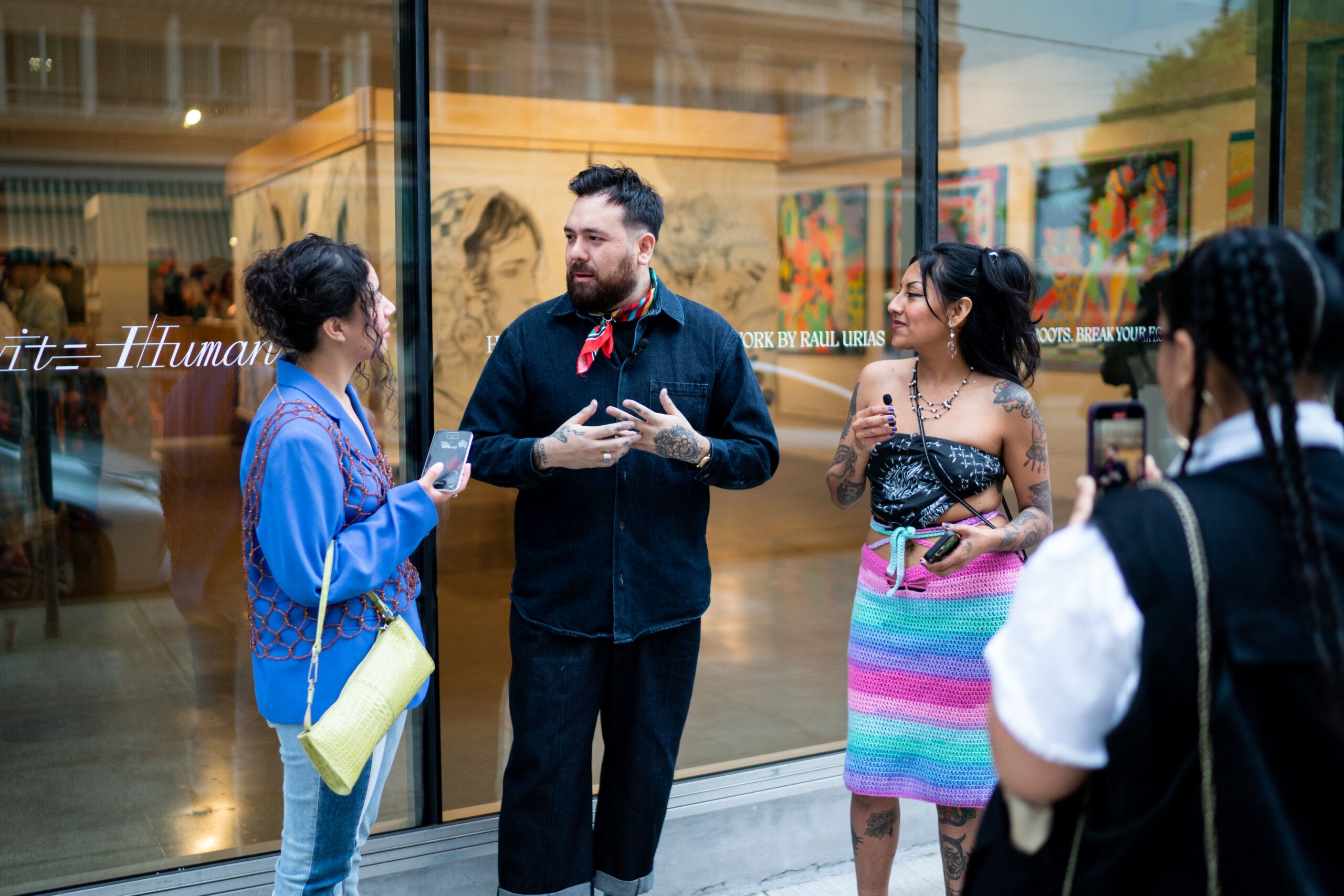 A group of people converse outside an art gallery. One person in the center is wearing a black outfit with a red scarf. Two individuals hold smartphones, and artwork is visible through the gallery windows.