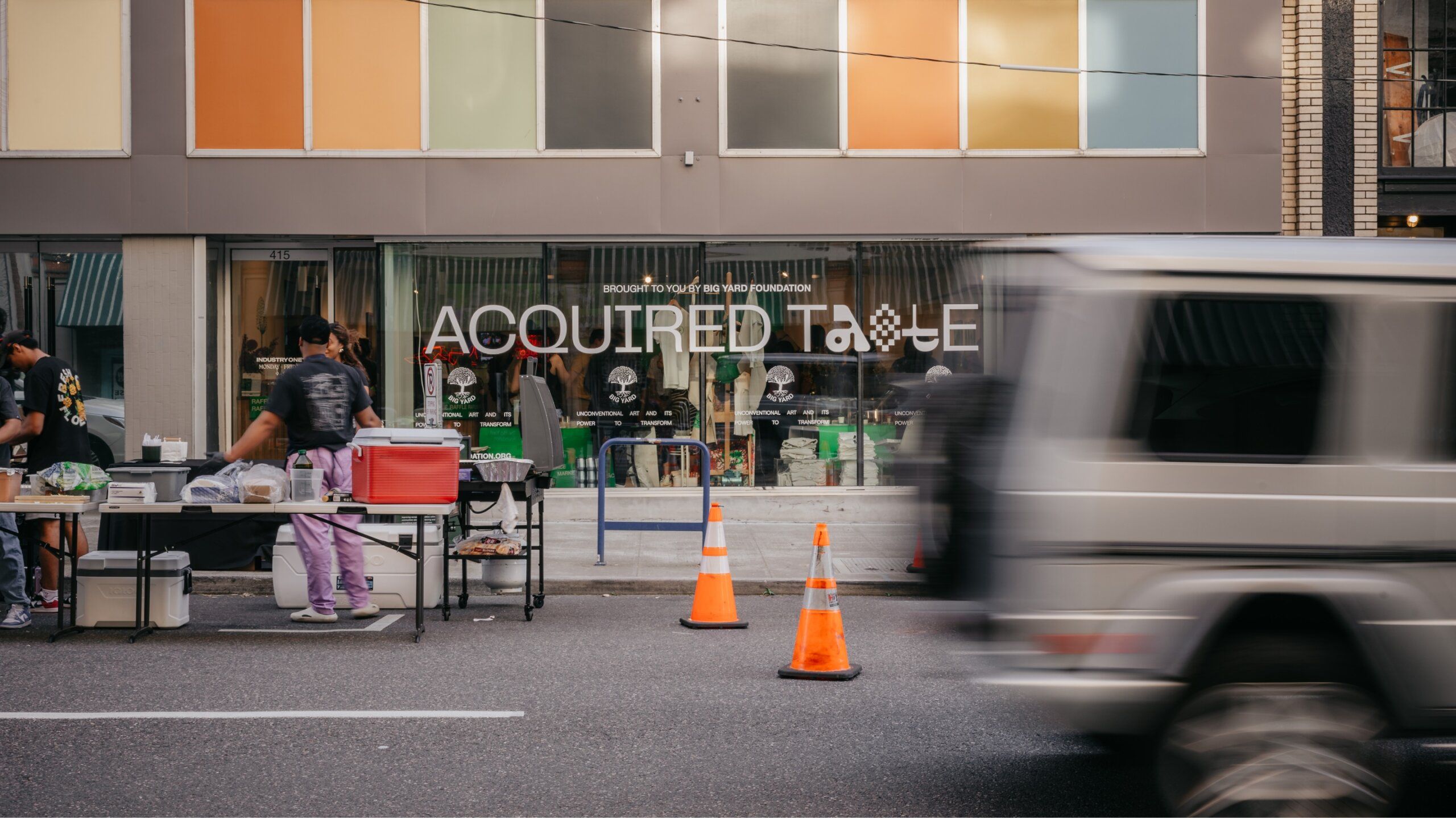 A street scene showing people at tables with various items outside a storefront. A moving vehicle and orange traffic cones are in the foreground. The storefront sign reads "ACQUIRED TASTE.