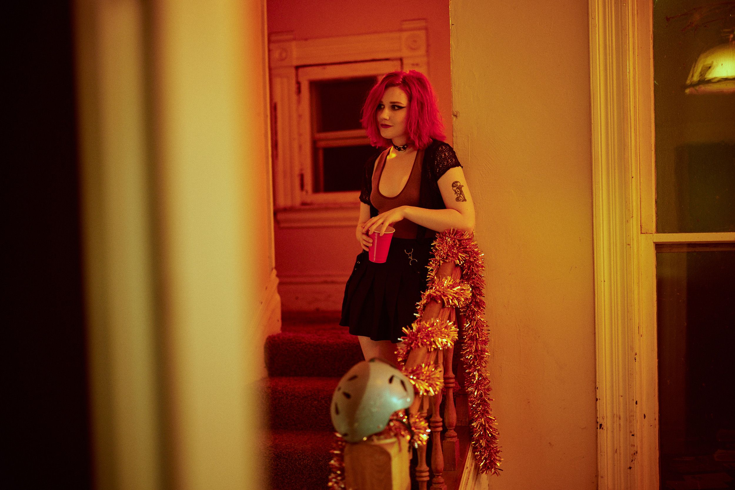 Person with red hair standing on stairs holding a red cup, next to festive tinsel and a railing with a helmet. Warm lighting.