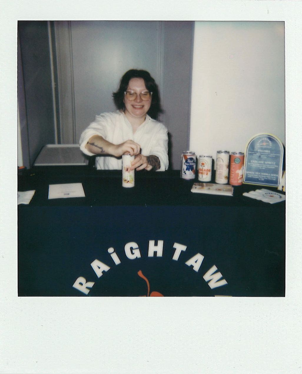 A person stands behind a booth with a dark blue table cover displaying "RAIGHTAW." They are opening a can, and there are multiple beverage cans and a plaque on the table.