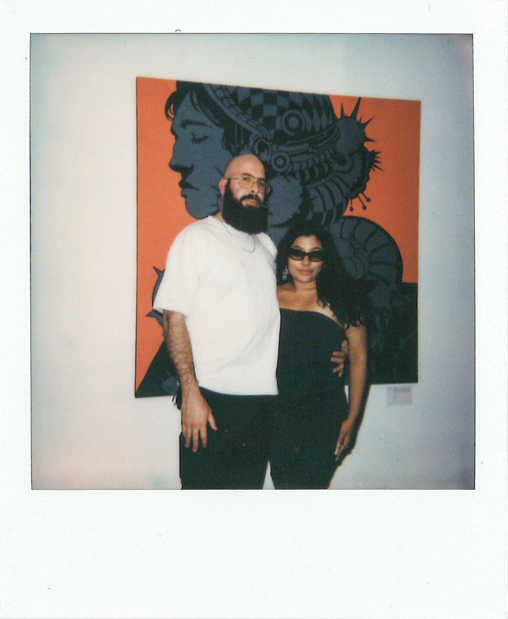 A man and woman standing together in front of a large artwork featuring a stylized profile of a person's head against a red background. The man has a beard and wears glasses, and the woman is wearing sunglasses.