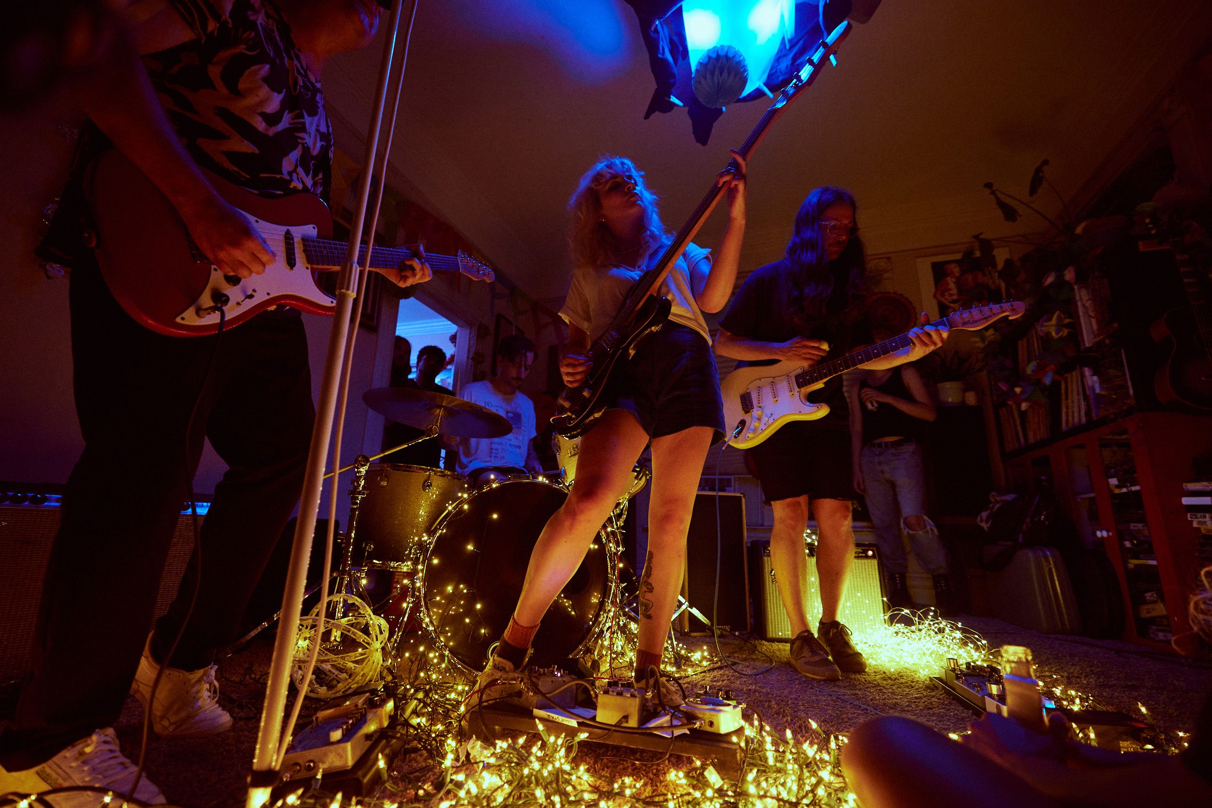 A band performs in a dimly lit room with string lights. The guitarist in the center plays energetically, surrounded by bandmates on guitar and drums.