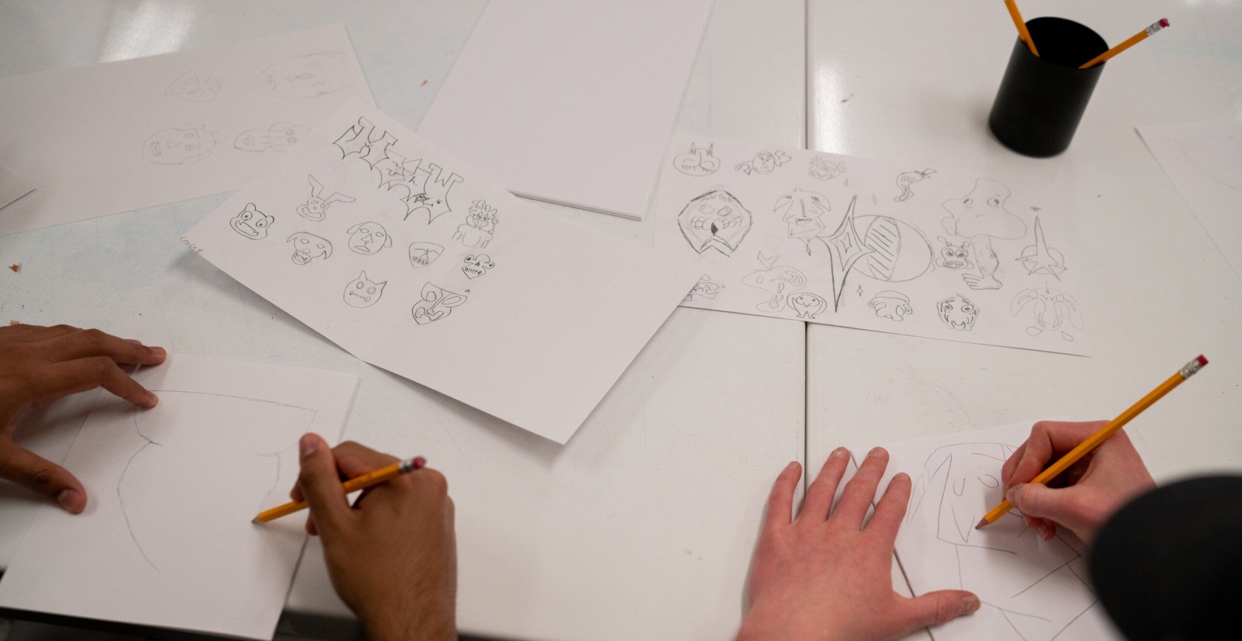 Two people sketching cartoon characters on white paper at a desk, with various drawings and pencils visible.
