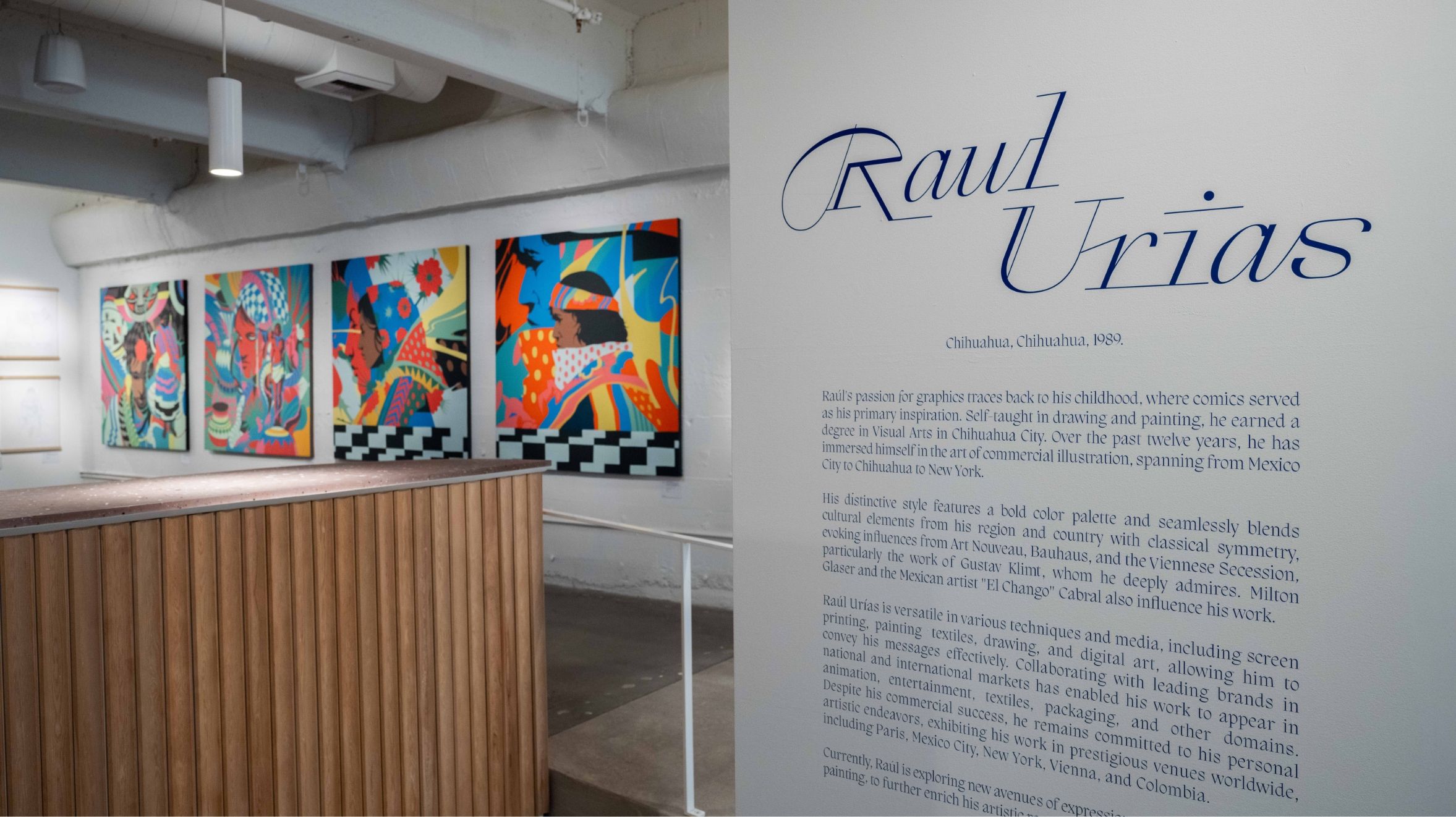 An art gallery displaying vibrant paintings by Raul Urias with an informational wall text about the artist on the right side.