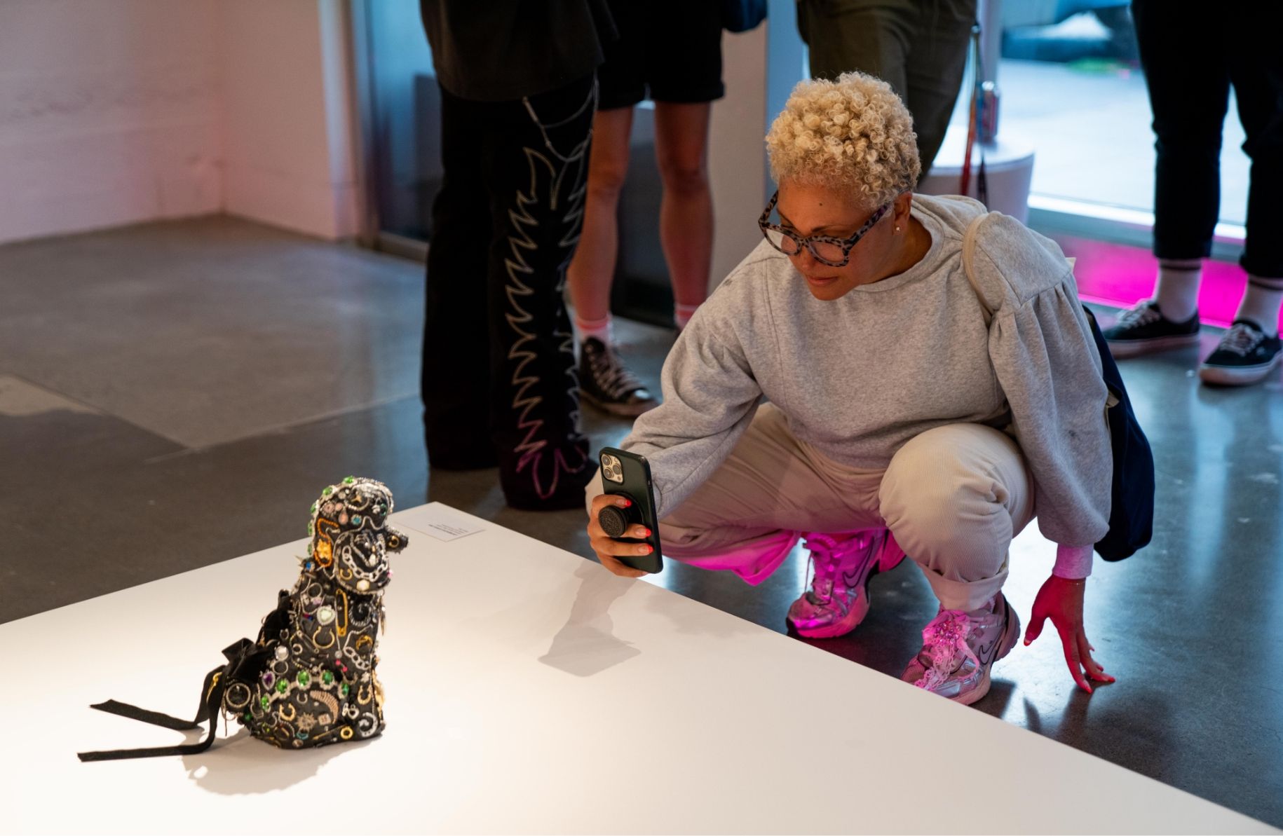 A person crouches to take a photo of a small, ornate sculpture on display in a gallery, surrounded by other visitors.