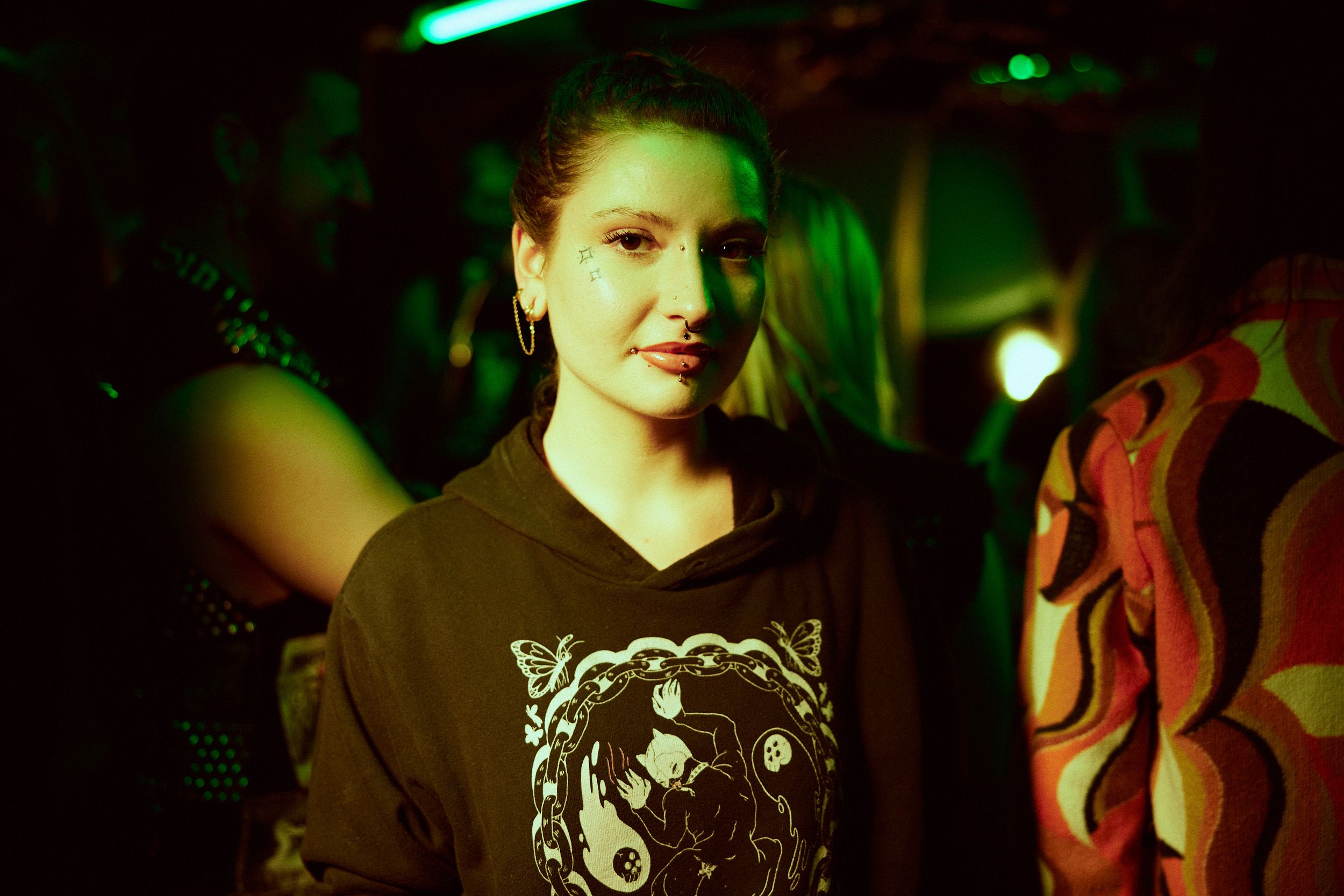 A woman in a dark hoodie stands in a dimly lit room with green lighting. She is surrounded by people and wears hoop earrings and facial piercings.