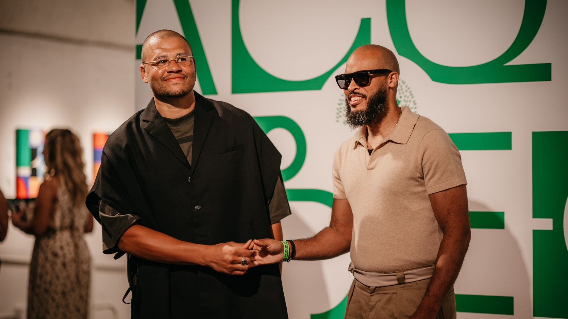 Two men shaking hands in front of a green and white wall at an indoor art gallery event. They are both wearing sunglasses and casual attire.