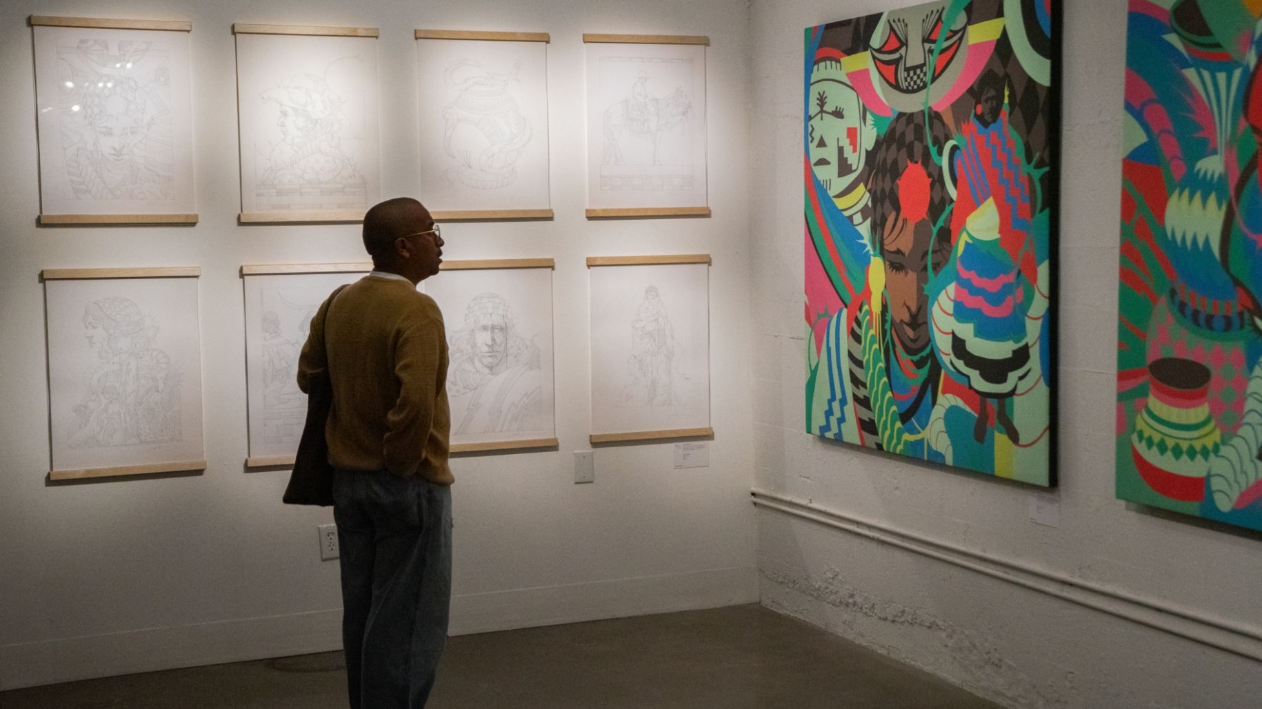 A person wearing a brown top and blue pants is observing a vibrant, multicolored painting in an art gallery. The wall to the left displays several sketches in frames.