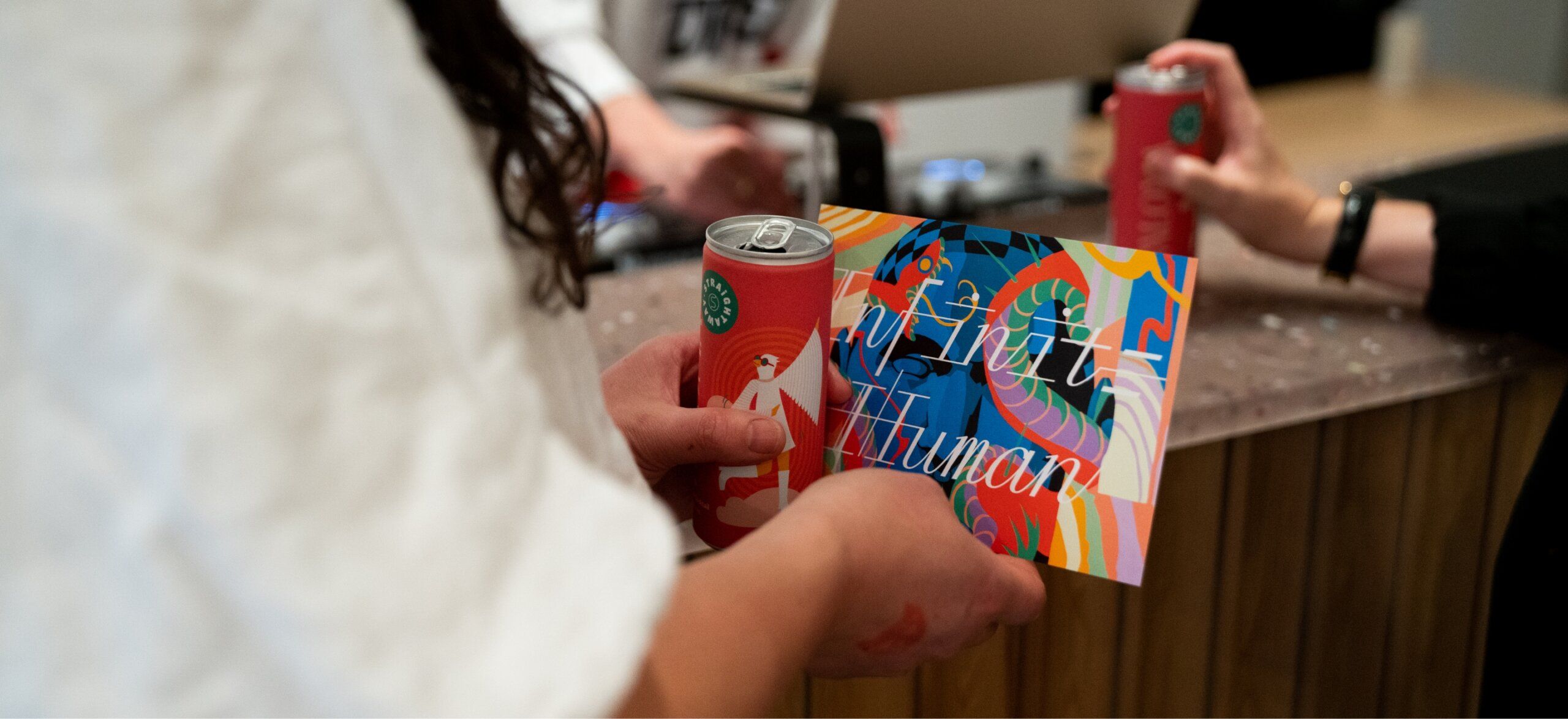 Person holding a colorful card and a can, while others are holding cans in the background. The card has text and a vibrant design.