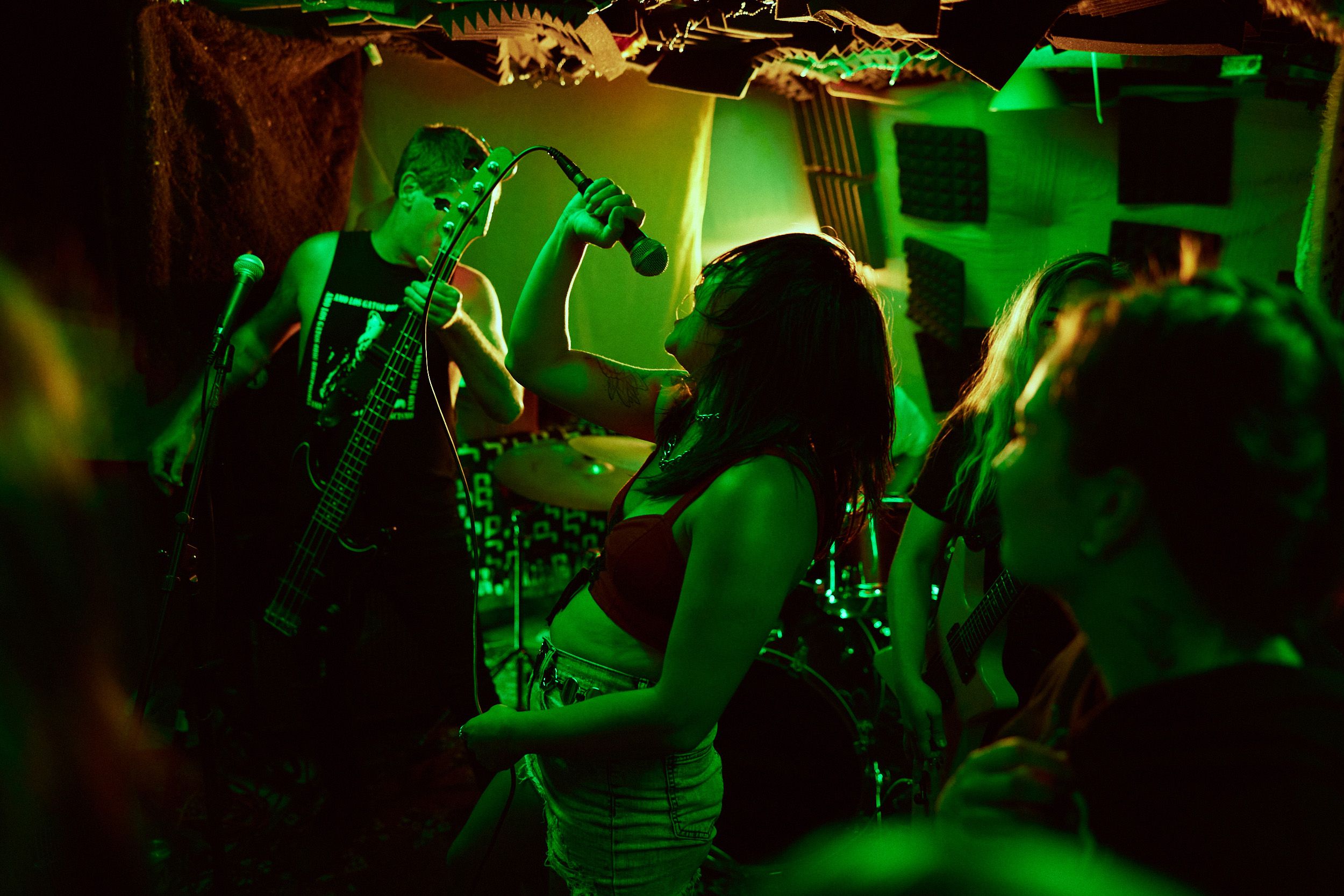 A band performs energetically in a dimly lit room with green lighting. The singer holds a microphone passionately, while band members play instruments in the background.