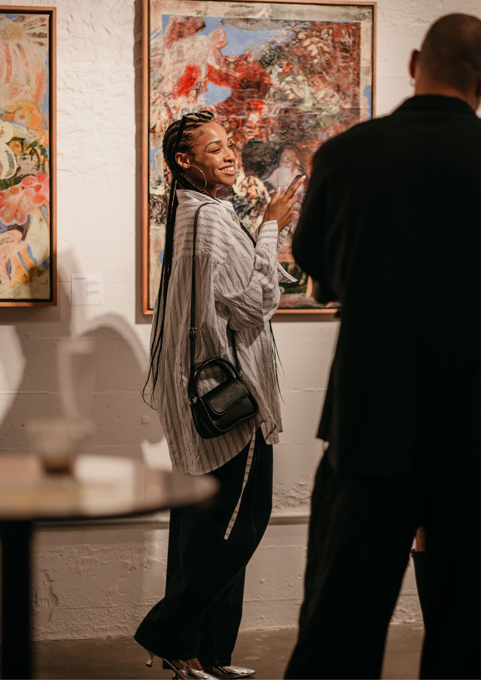 A person with long braids, wearing a striped shirt and holding a small black bag, smiles and gestures while looking at art in a gallery.