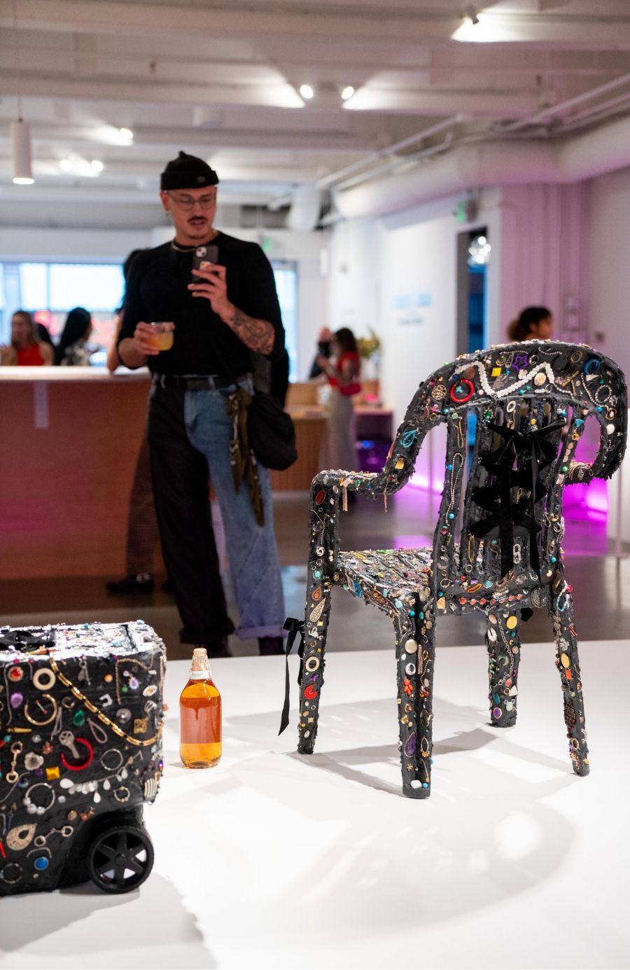 A person taking a photo in a gallery with a decorated chair and a textured box on display.