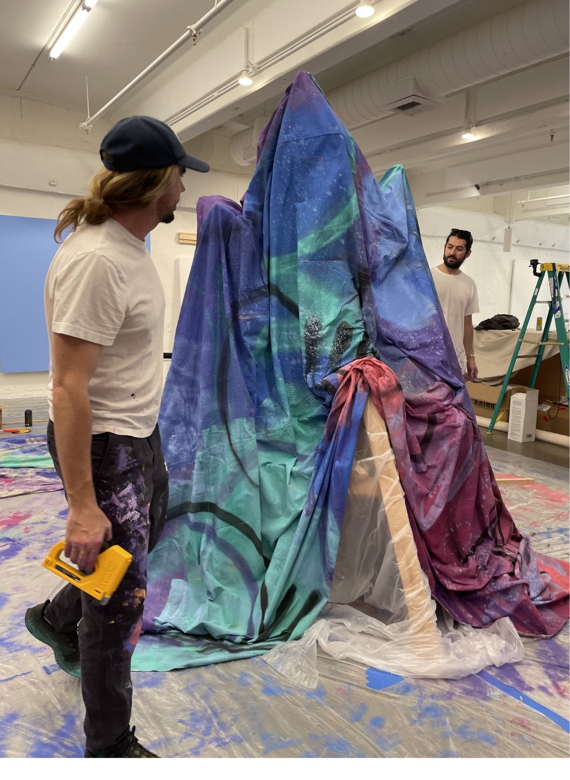Madsteez stands in front of painted tarp sculpture. He is holding a staple gun. Its a living stands behind sculpture