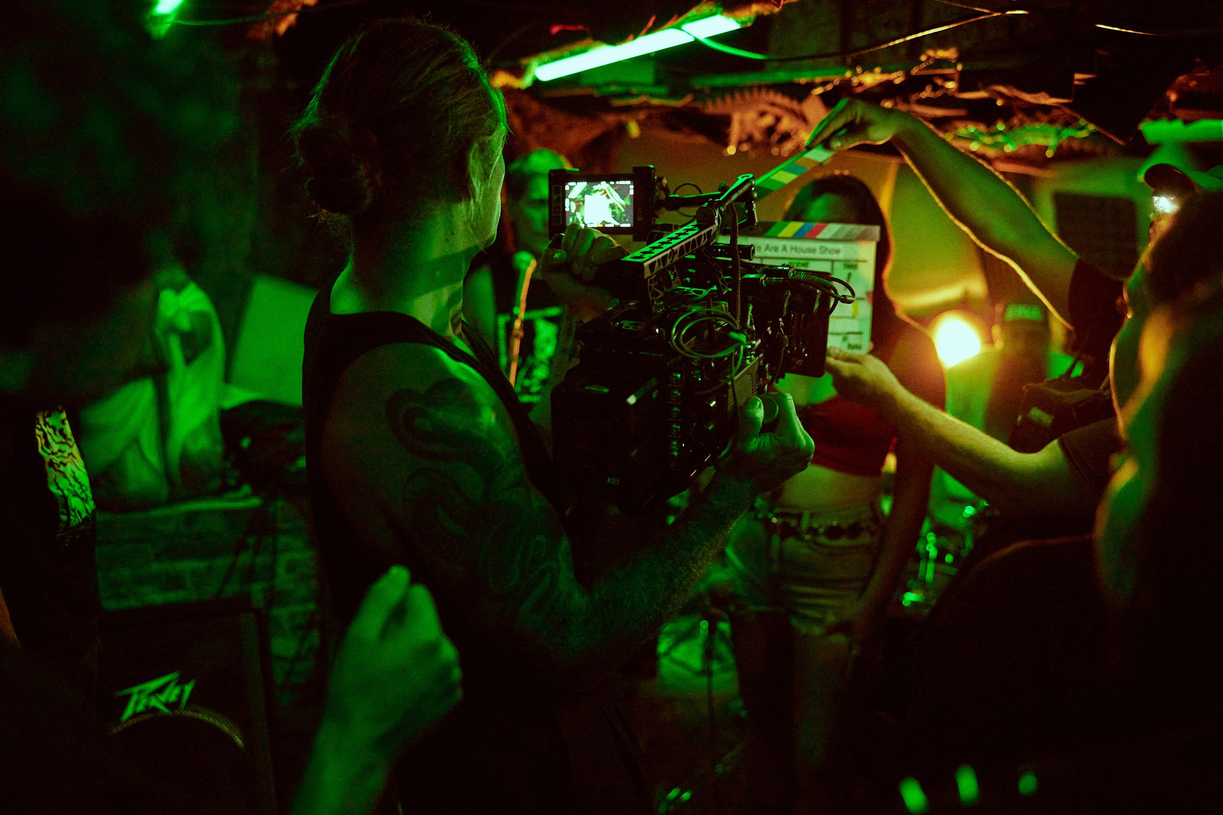 A camera operator films a scene with a clapperboard in a dimly lit, green-tinged environment. People are gathered around the scene.