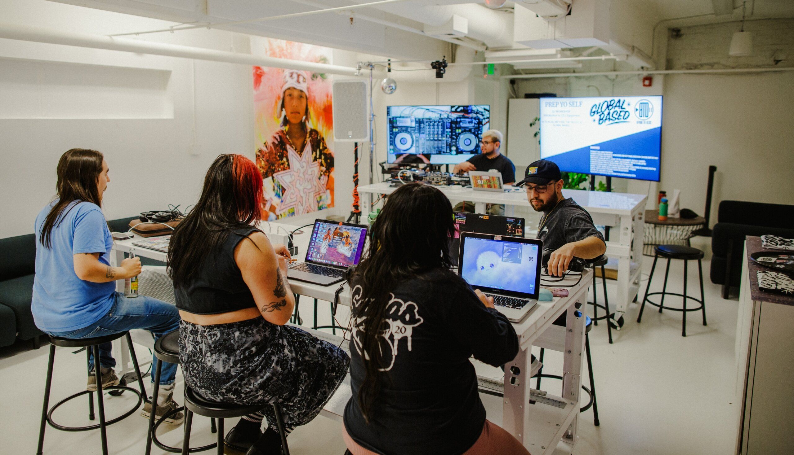 Several people work on laptops at a long table in a room with a large screen displaying digital art and text.