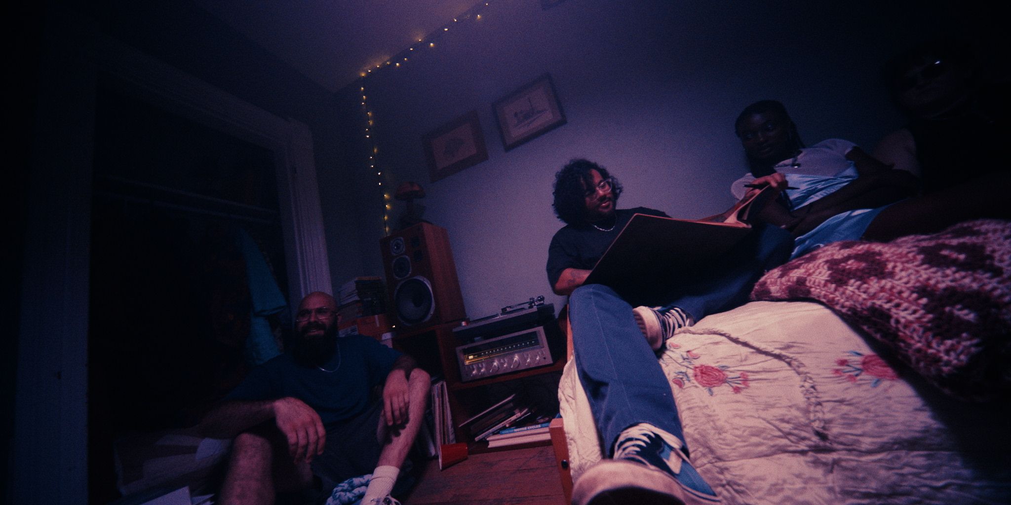 A group of people sitting in a dimly lit room with soft lighting. One person sits on the bed holding a book. Speakers and framed pictures are visible in the background.