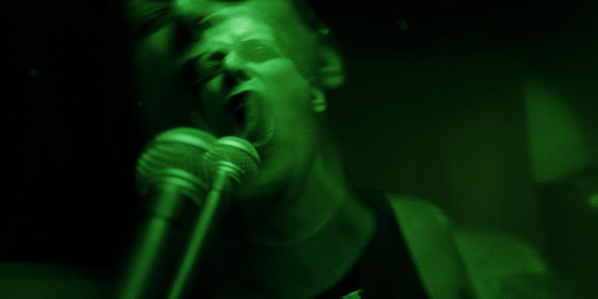 A person passionately singing or shouting into two microphones, with a green filter and motion blur effect.