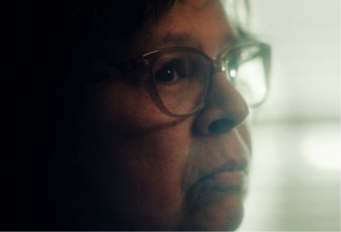 Close-up of a person wearing glasses, looking serious, with soft lighting from the side. Background is blurred.