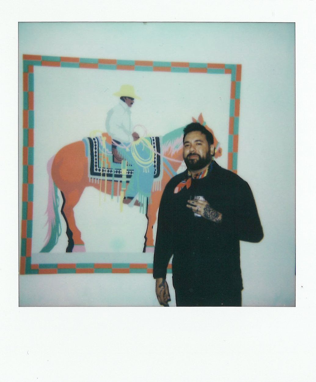 A bearded man with tattoos stands in front of a colorful painting depicting a person on a horse.