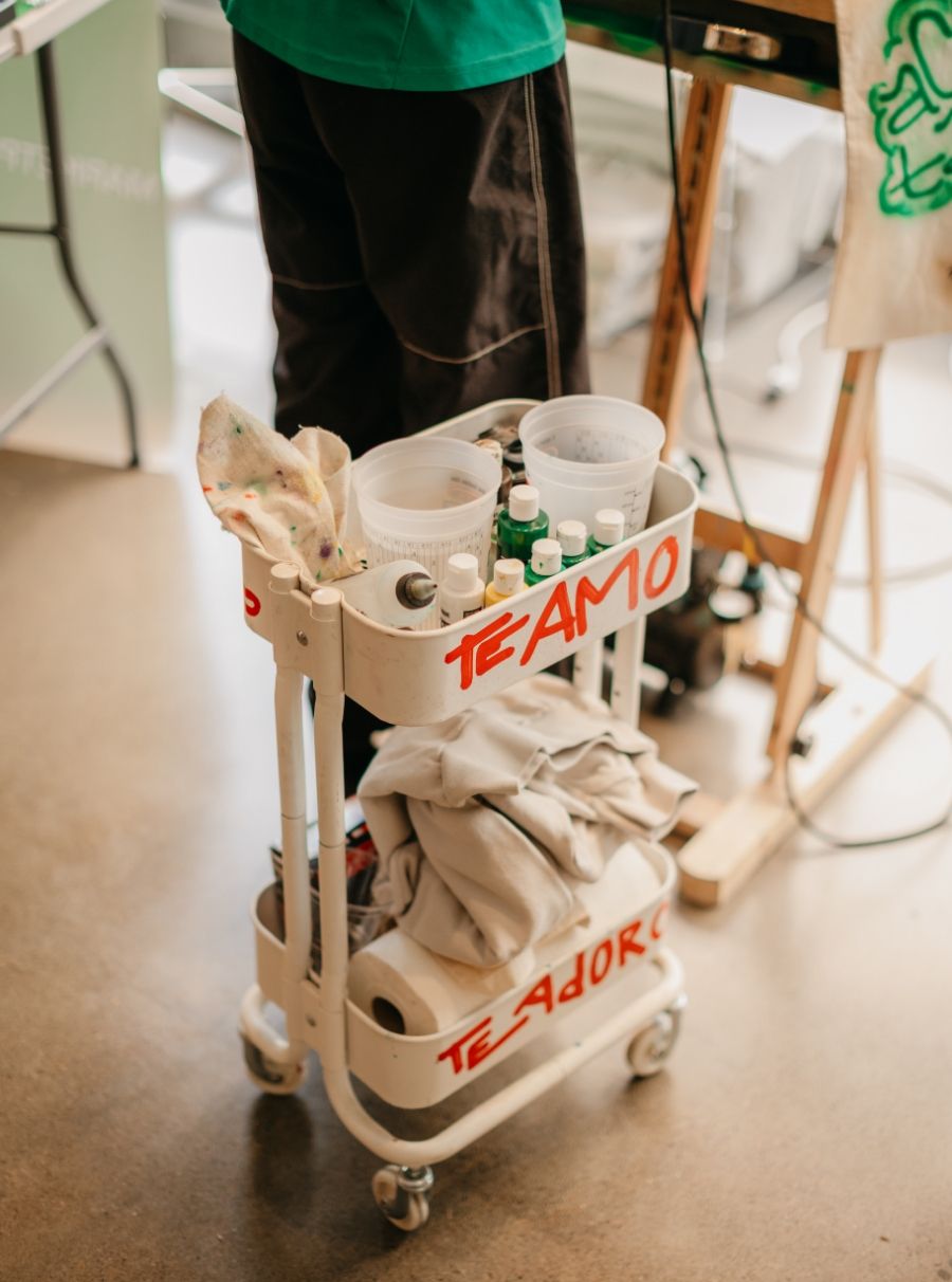 A wheeled cart holds various art supplies, including bottles, cloths, and containers marked with the words "TE AMO" and "TE ADORO." In the background, a person in black pants is partially visible.