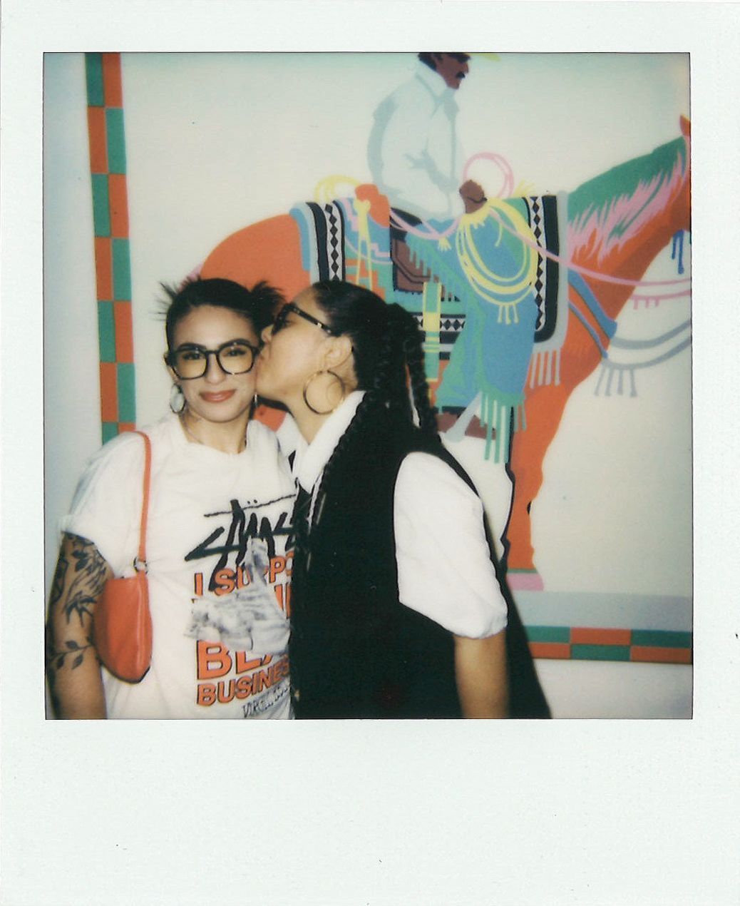 Two people stand in front of a colorful painting of a person on horseback. One person kisses the other on the cheek while both smile. The person getting kissed has tattoos and wears glasses.