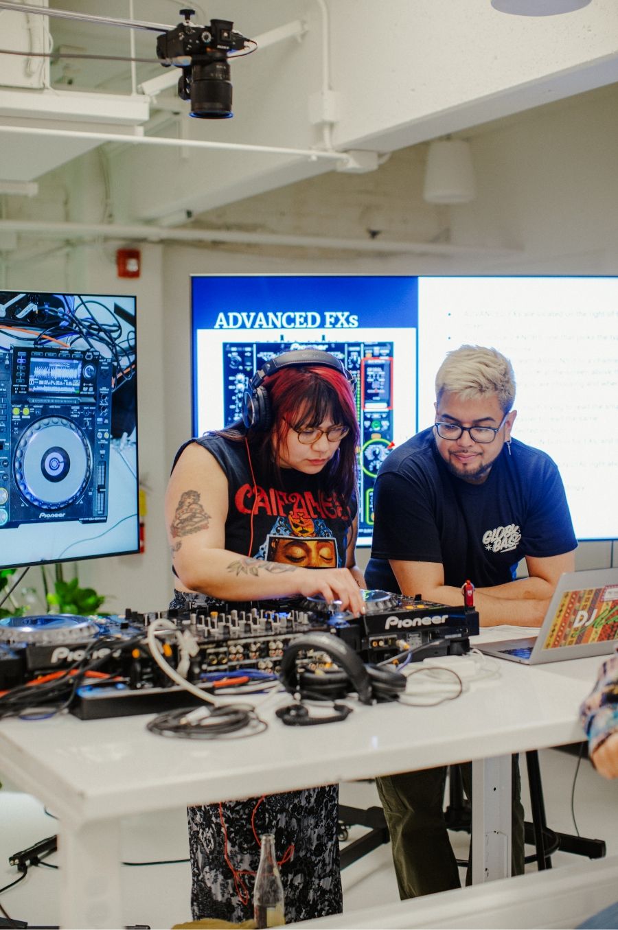 Two individuals are working together in a modern studio setting with audio equipment. One is adjusting controls on a DJ mixer, while the other looks at a laptop. Behind them, screens display advanced sound effects.