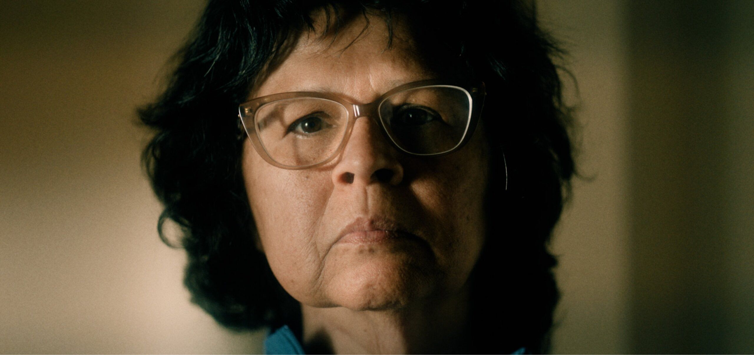 A person with glasses and short, dark hair looks ahead with a serious expression, set against a softly blurred background.