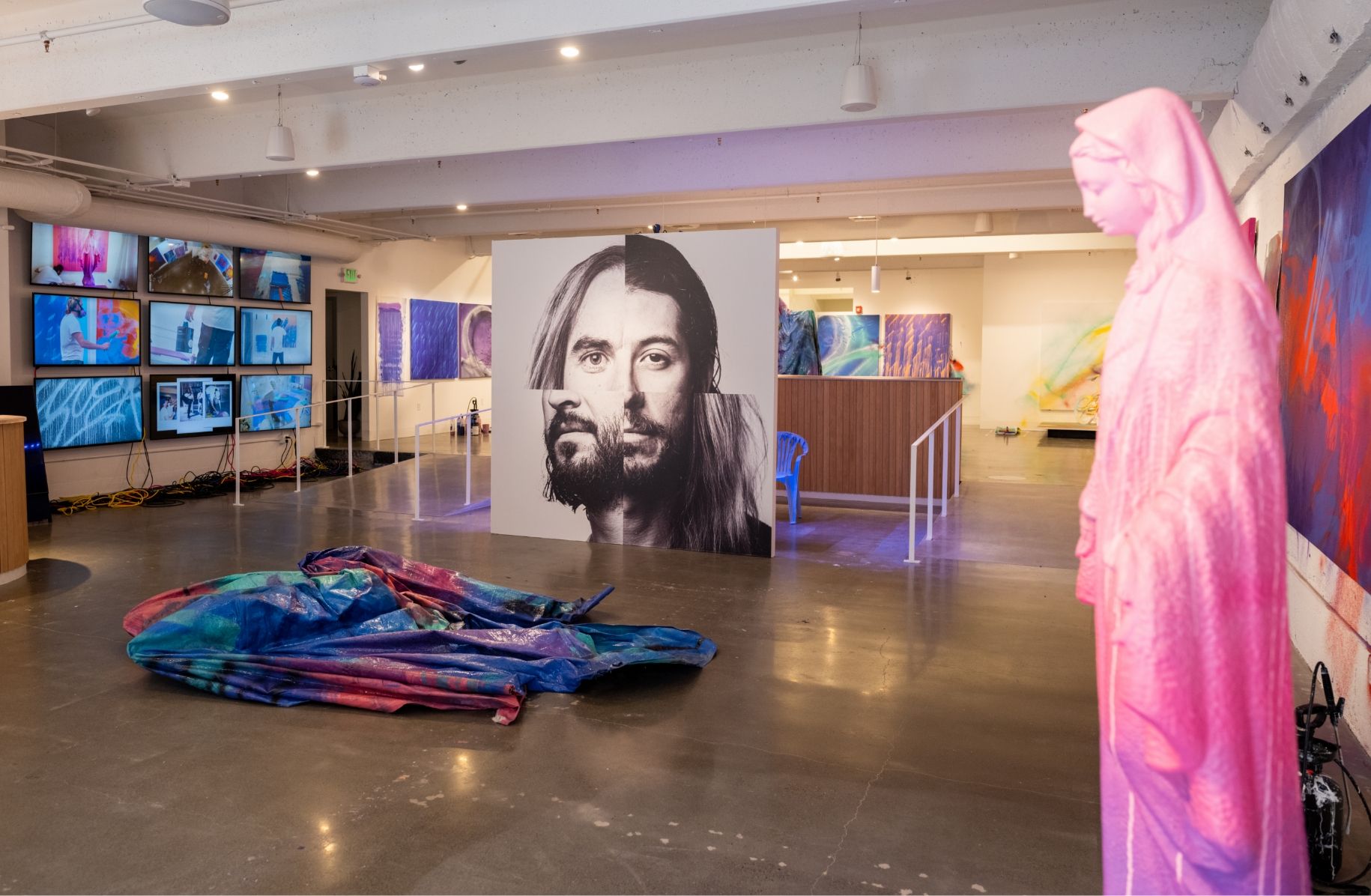 View of whole show from gallery floor. Mother Mary spray painted statue, painted tarp sculpture, face montage and video screen wall prominent