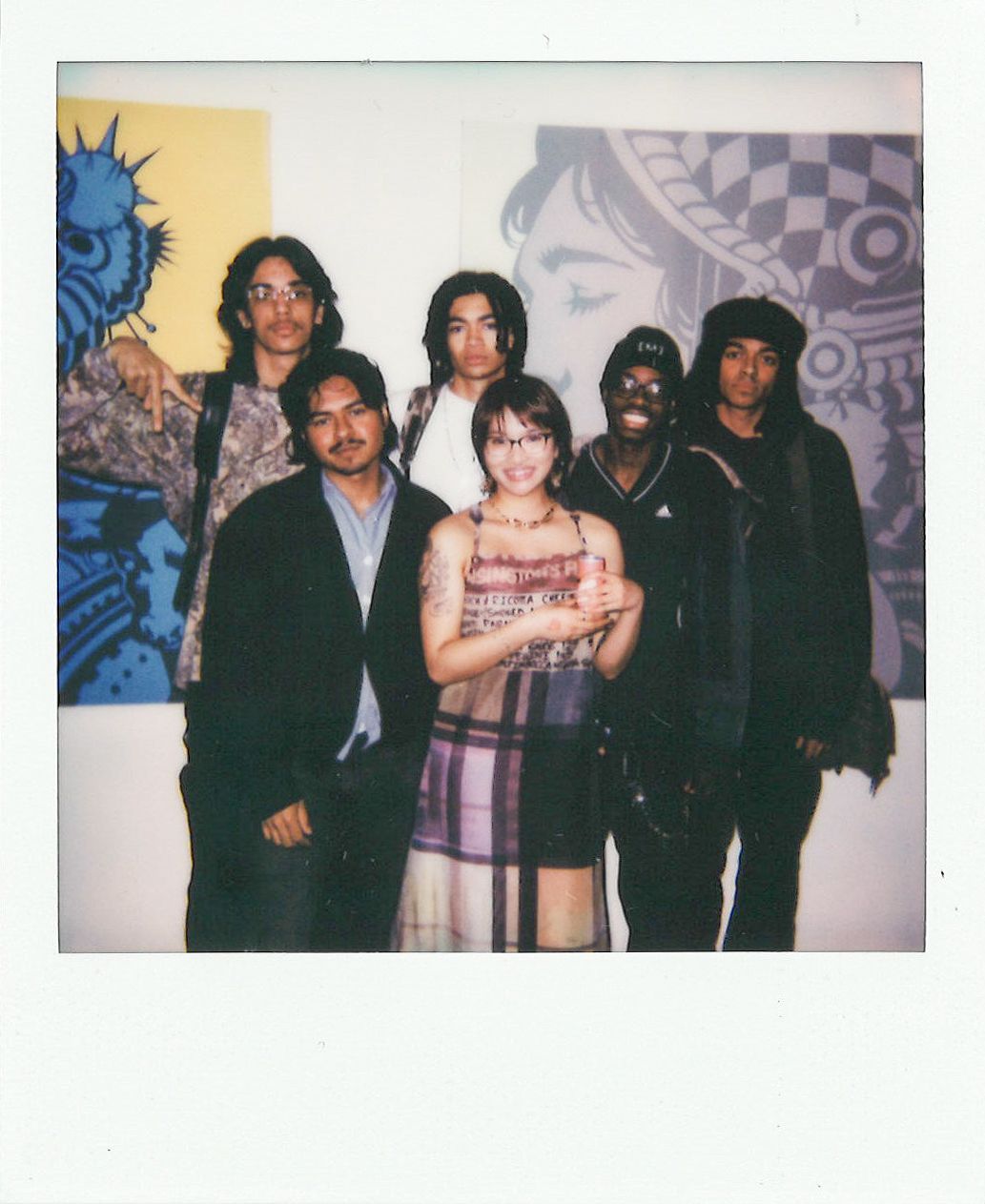 A group of six people posing in front of colorful artwork, with one person holding a cat and everyone dressed in casual, eclectic clothing.