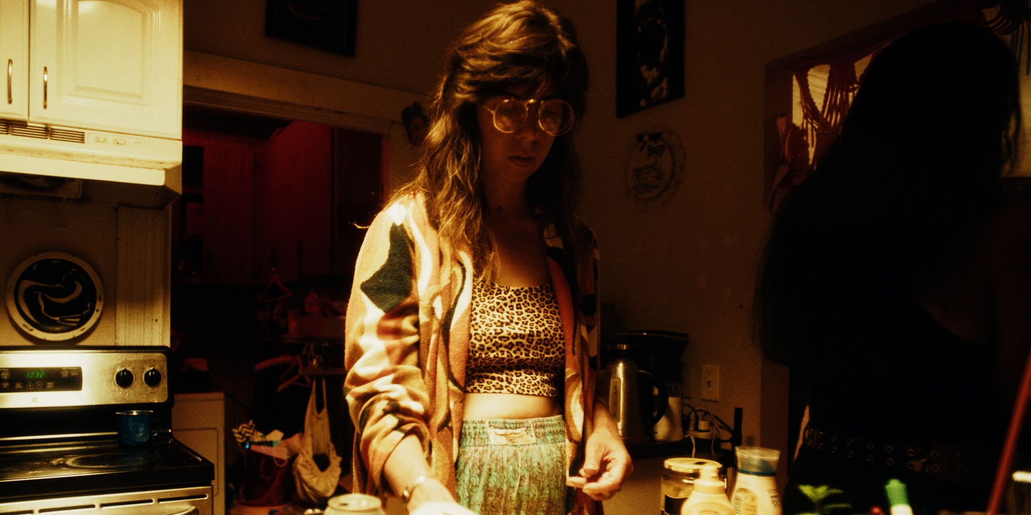 A woman in a dimly lit kitchen stands near a countertop, wearing glasses and a patterned top.