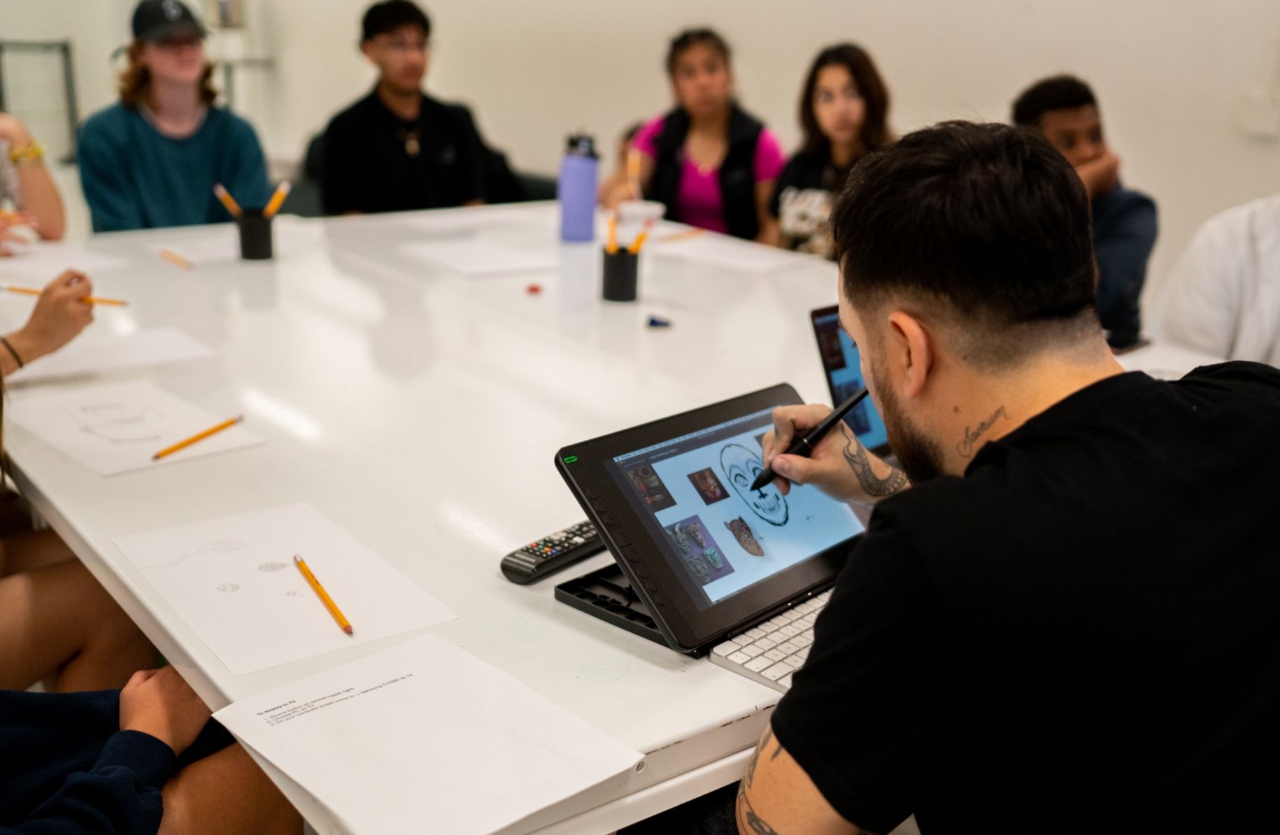 Man working on a laptop with digital artwork displayed on the screen, surrounded by attentive students seated around a long white table in a classroom.