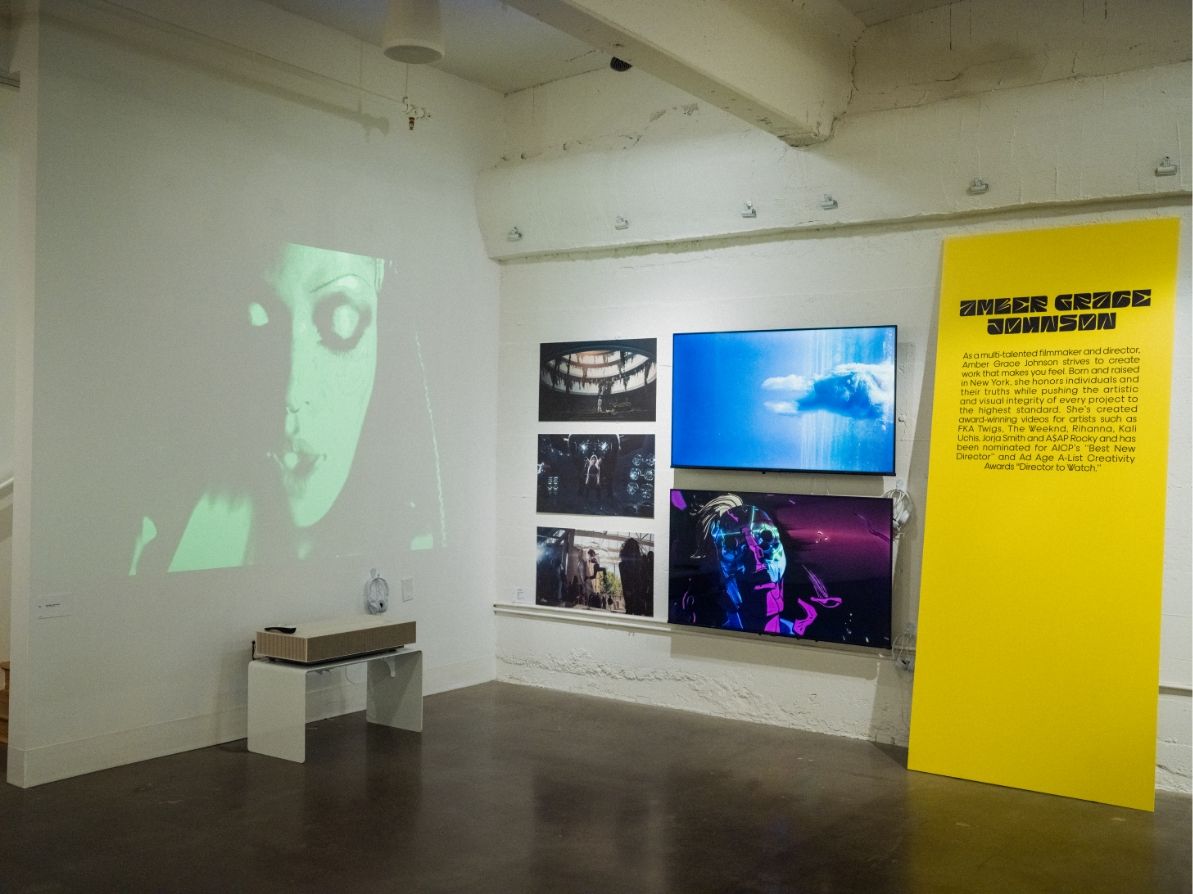 Corner of Gallery showing Amer Grace Johnson's work including a projection of a video, 2 TV's, 3 video still prints and a large yellow board with her bio