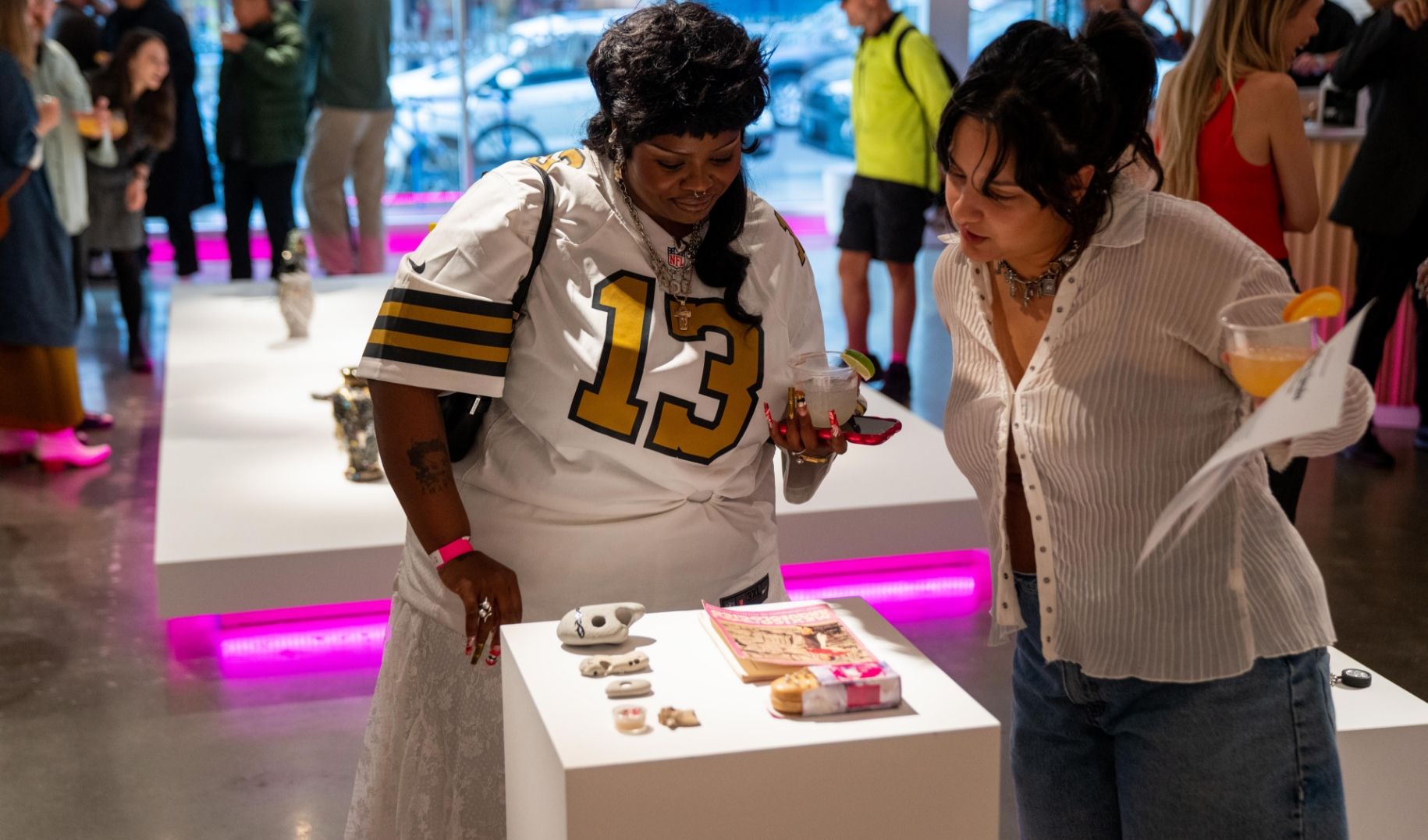 Two people examining small objects on a display table in a bright gallery environment. Other people are seen in the background.