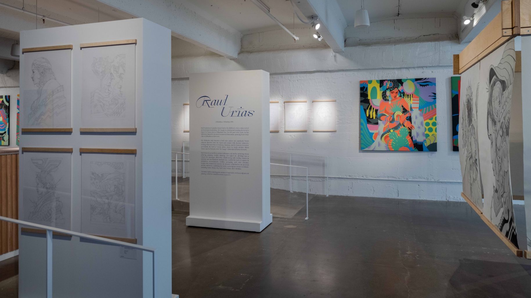 An art gallery featuring sketches and colorful abstract paintings on white walls. Text panel in the center provides artist information. Lighting illuminates the artwork in an otherwise dim space.