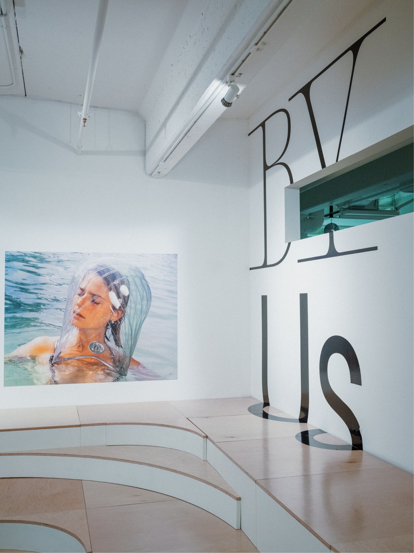 Rear corner of gallery showing stadium seating, video still print of woman floating in water with head in clear covering. Words By Us in vinyl on wall