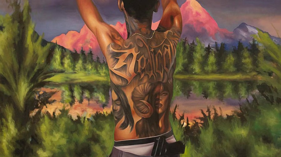 A person with extensive back tattoos, including a ram and a person's face, stands in front of a scenic mountain landscape at sunset with their arms raised and a gun tucked into their waistband.