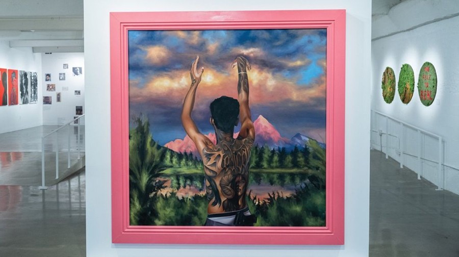 A framed painting depicts a shirtless person with tattoos, raising arms towards a colorful sky with mountains and trees in the background, displayed in an art gallery.